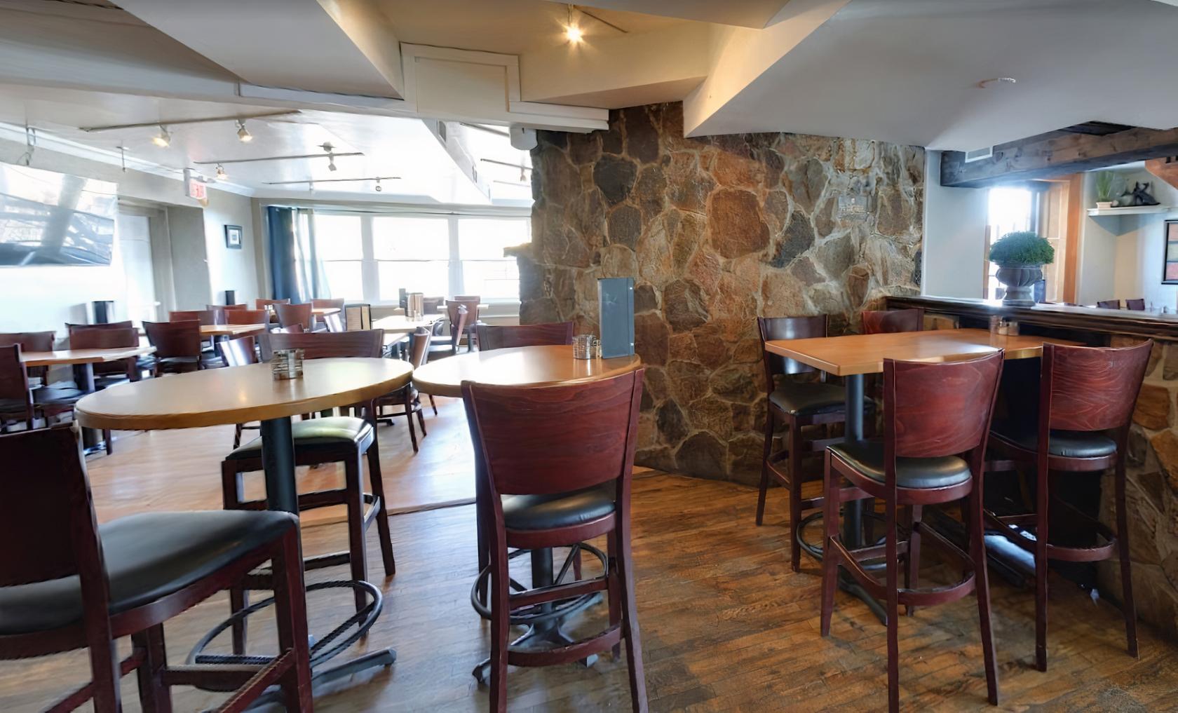An event space in a pub with round wooden tables, high-backed chairs, and a stone accent wall at The Olde Stone Cottage Pub.