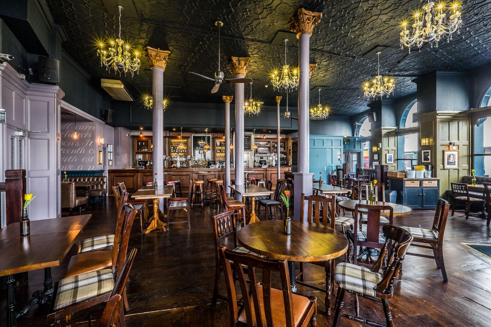 A pub with ornate chandeliers, wooden tables and chairs, decorative columns, and a classic bar at The Albany.
