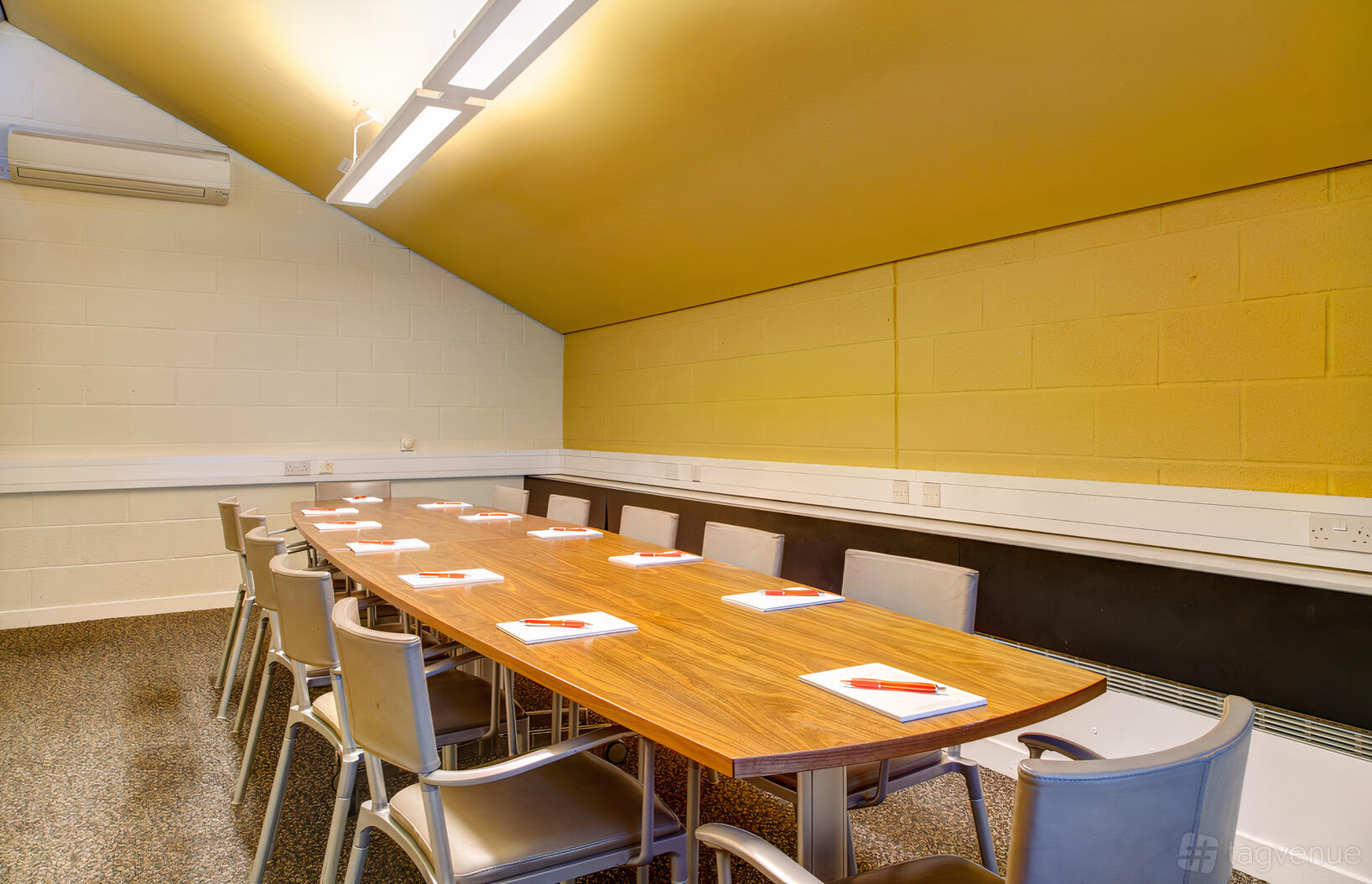 A meeting room with a long wooden table, padded chairs, yellow sloped ceiling, and notepads at Sadler's Wells.