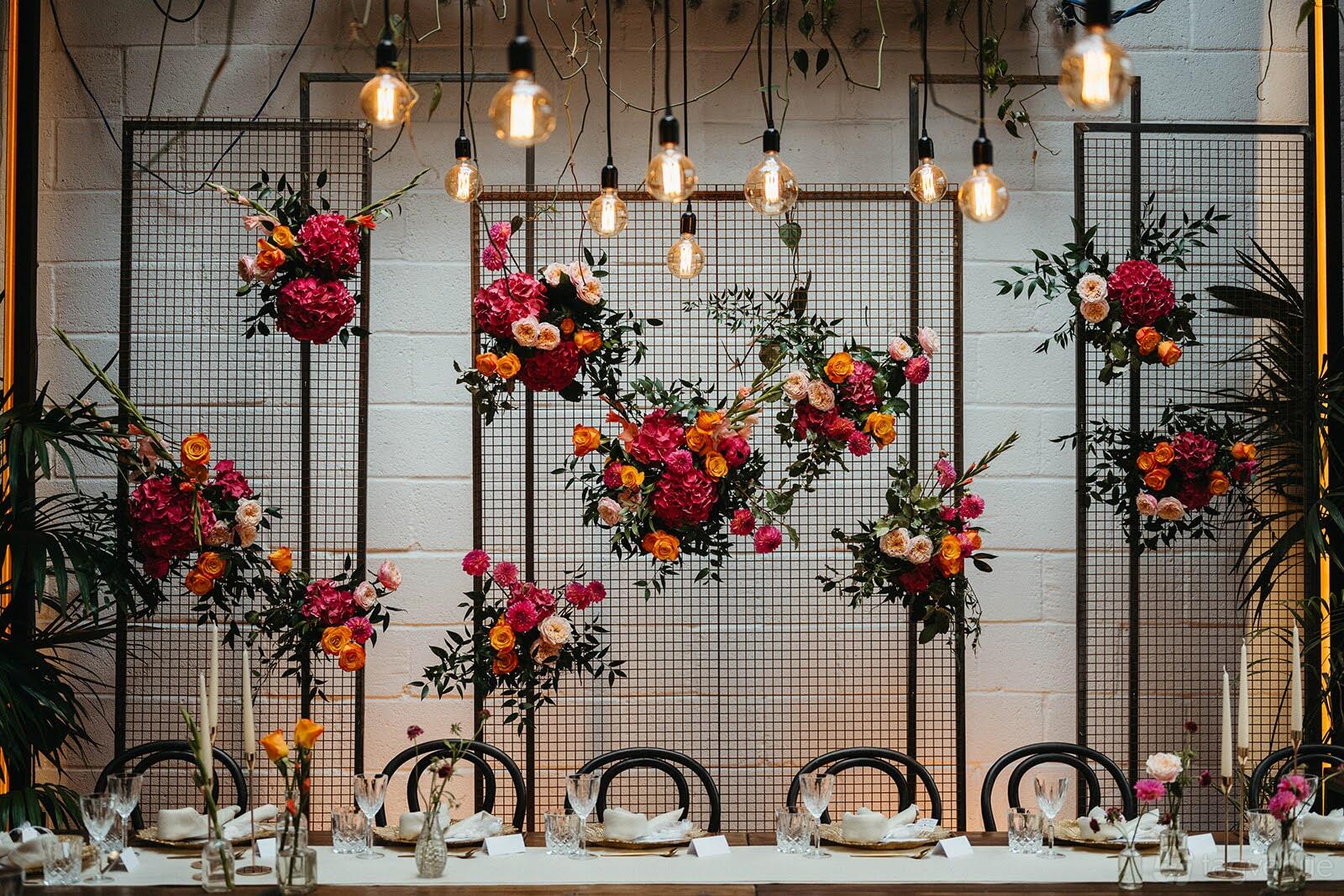A cafe with floral arrangements on metal screens, hanging Edison bulbs, and a long table set for dining at The Hackney.