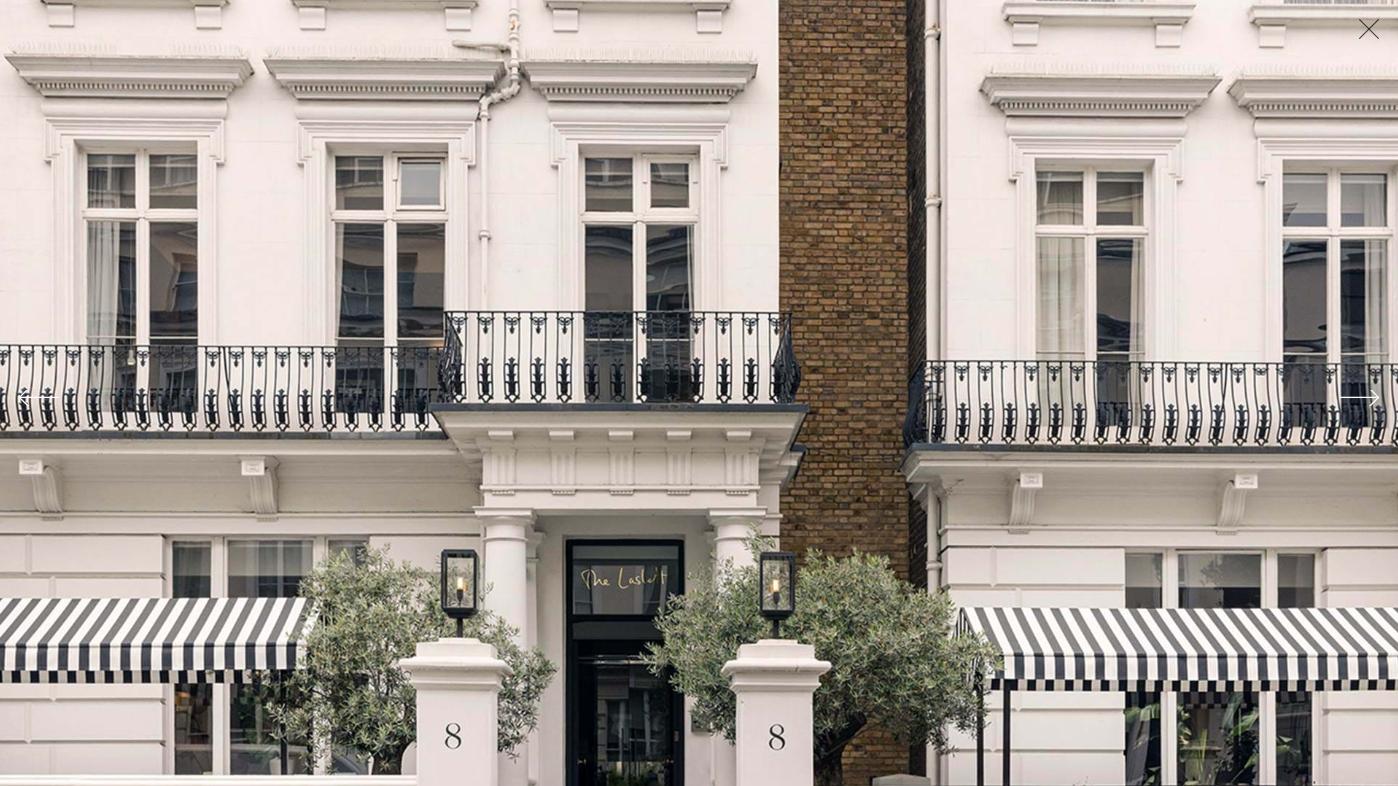 A hotel exterior with white townhouse façade, black railings, striped awnings, and potted trees at The Laslett.
