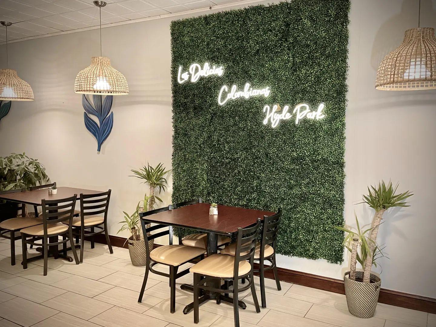 A restaurant interior with a faux greenery wall, neon sign, wooden tables, and wicker pendant lights at Delicias Colombianas Hyde Park.