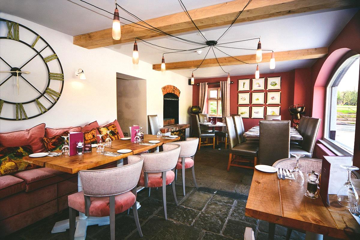 A pub dining room with exposed wooden beams, a large wall clock, rustic wooden tables, and upholstered seating at The Ashton.