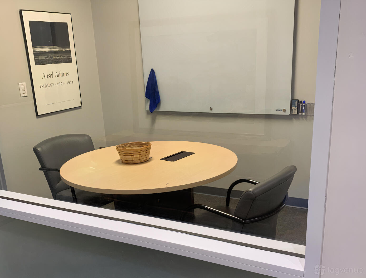 A meeting room with a small oval table, two black chairs, and a wall-mounted whiteboard at Origin Cowork.