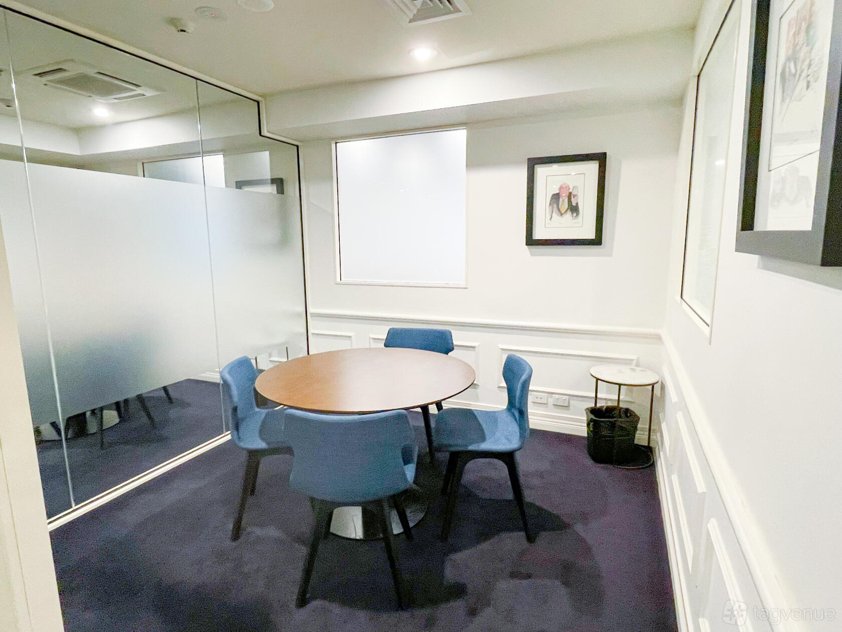 A meeting room with a round table, blue chairs, framed art, and frosted glass walls at Christie Spaces Conferencing Melbourne.