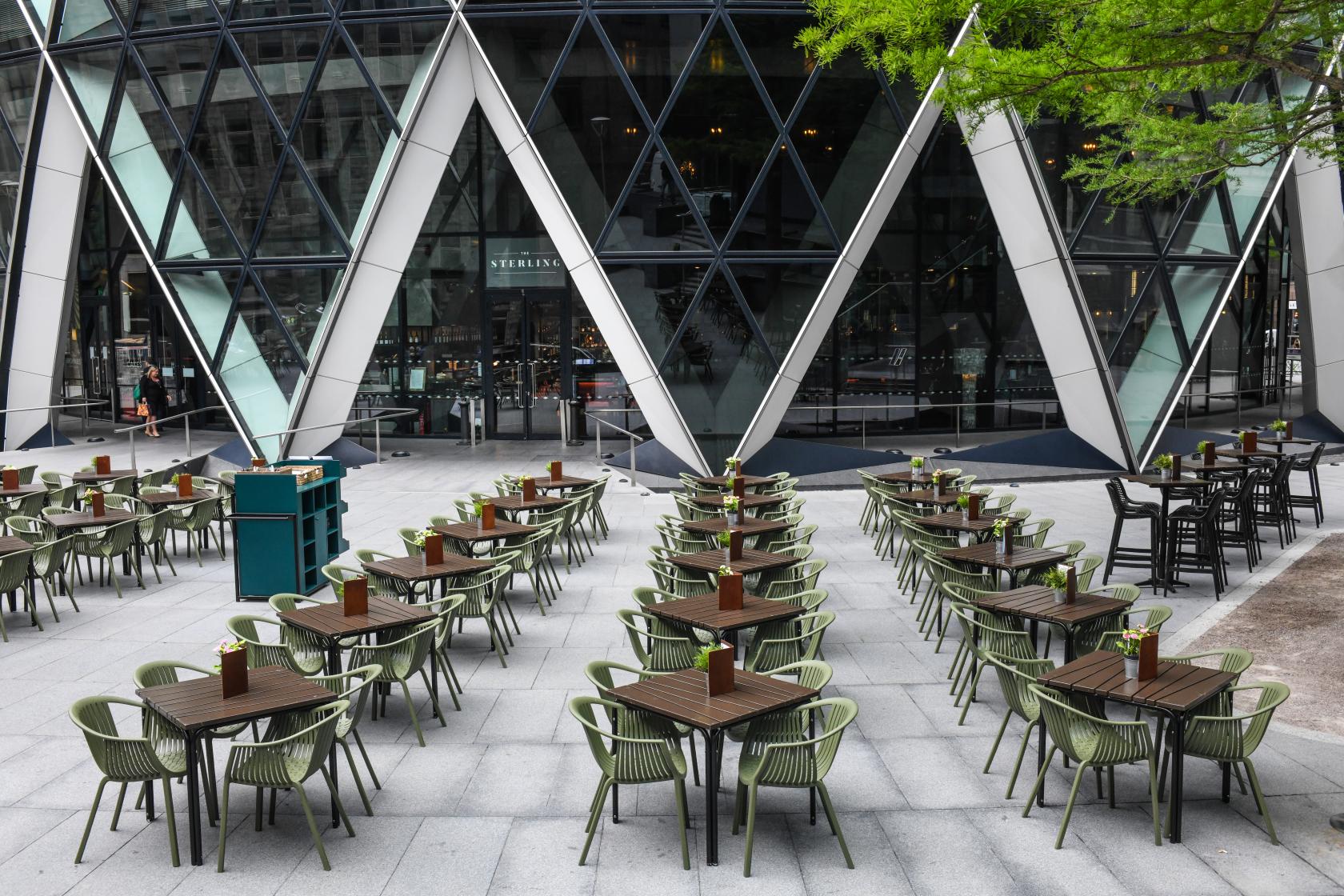 A restaurant terrace with rows of outdoor tables and green chairs overlooking modern glass architecture at The Sterling.
