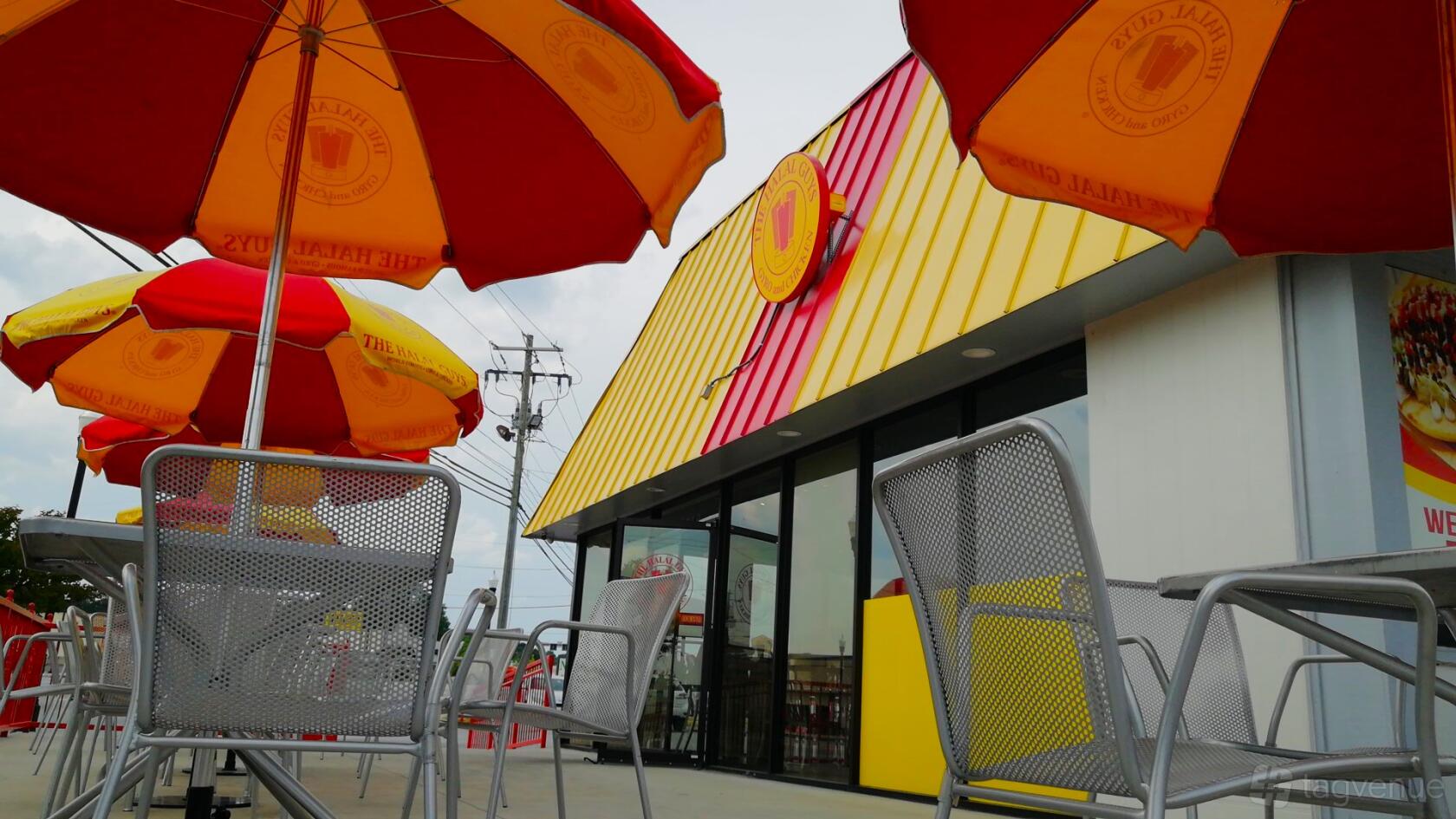 An outdoor dining area with metal patio chairs, tables, and red-and-yellow branded umbrellas at The Halal Guys.