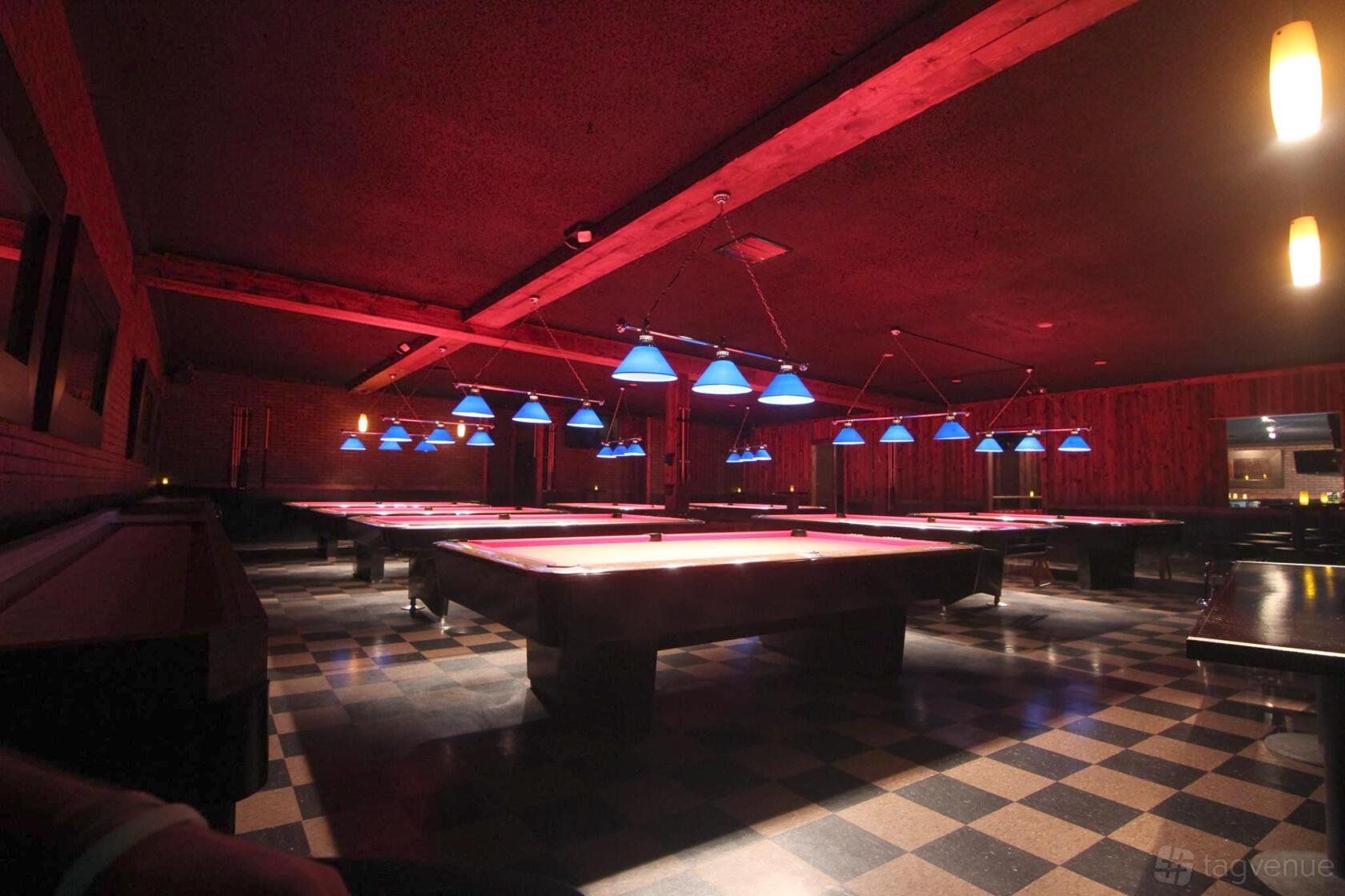 A bar with multiple pool tables under blue pendant lights on a checkered floor at The Brickyard Pub @ Hollywood.