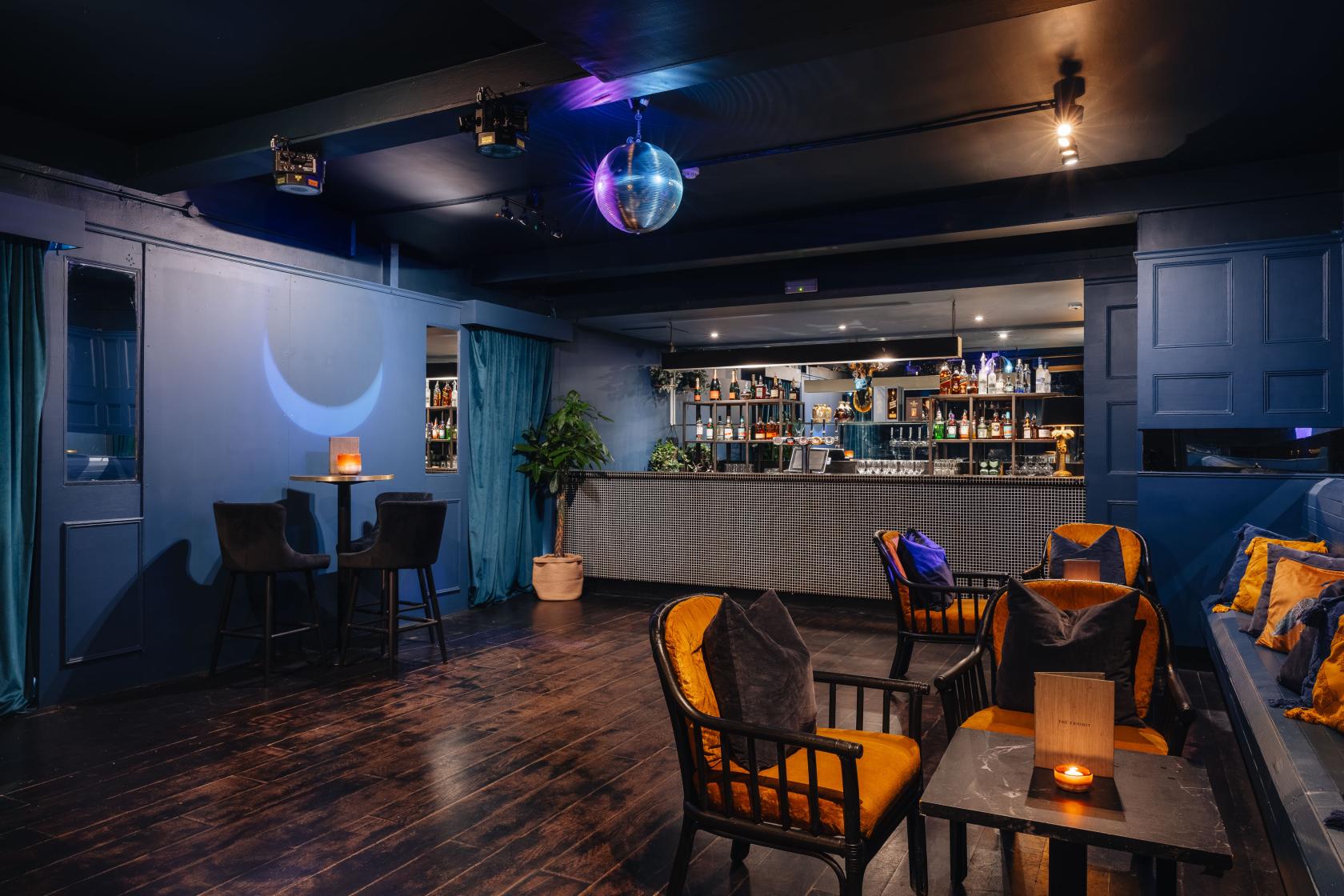 A function room in a cocktail bar with dark wood floors, velvet armchairs, and a mirrored disco ball at Exhibit.
