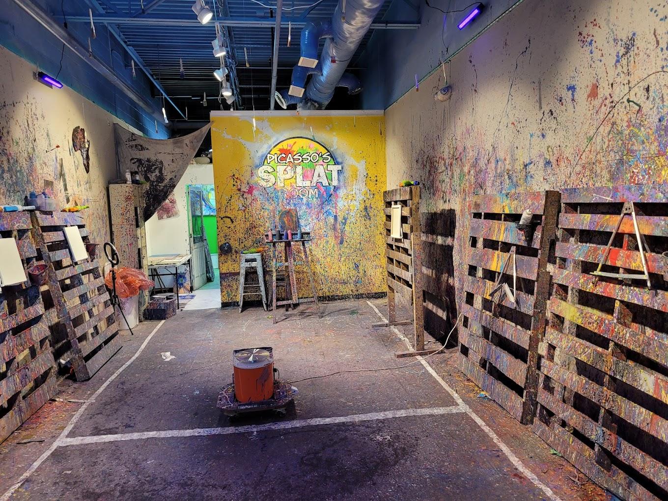 An art studio with paint-splattered walls, wood panel partitions, easels, and exposed ceiling pipes at Picasso's Splat Room.