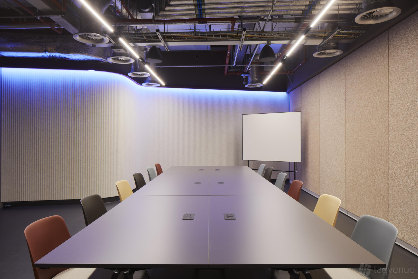 A meeting room with a long conference table, colorful chairs, acoustic wall panels, and exposed ceiling at Storey Club Broadgate.