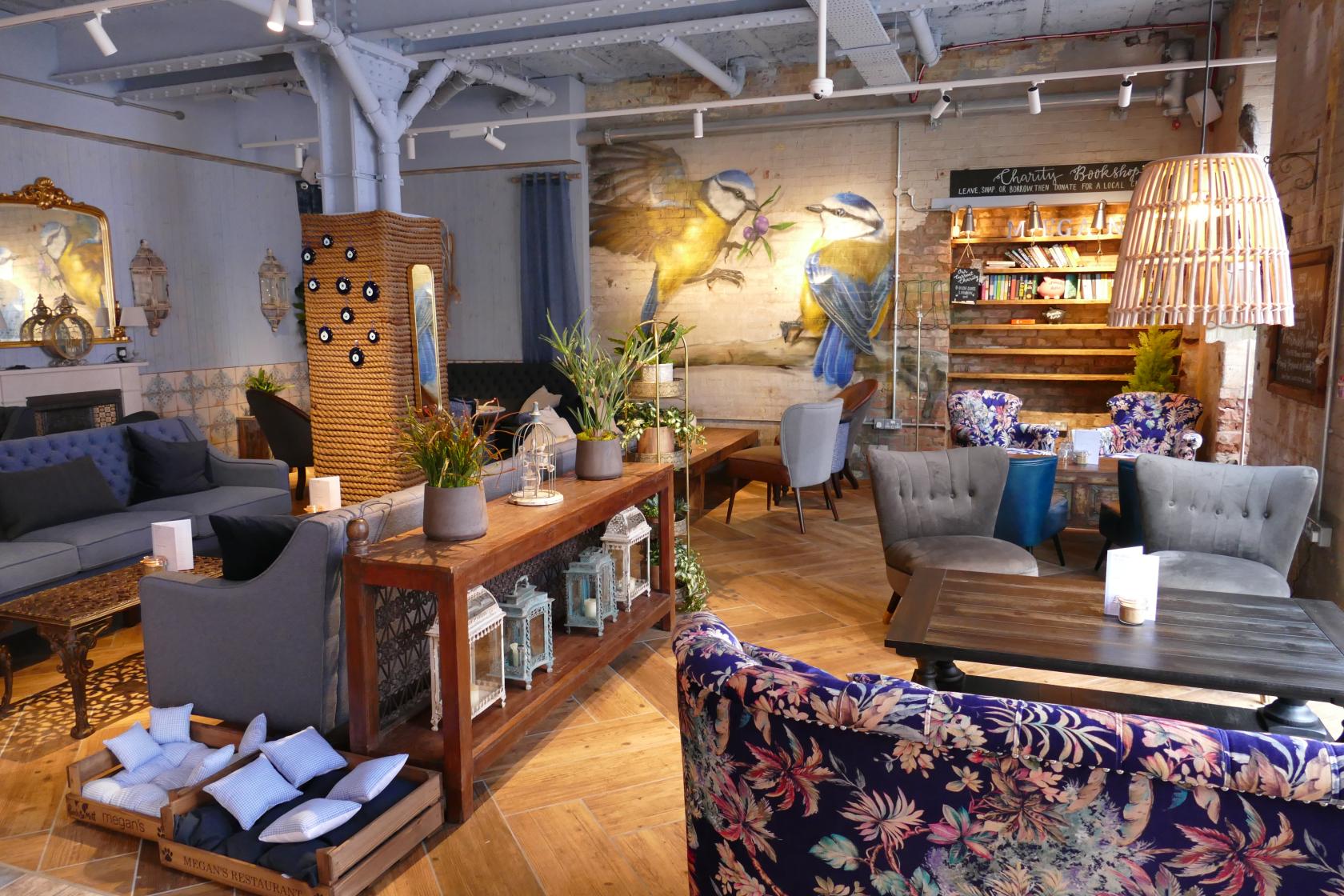 A restaurant lounge with patterned armchairs, wood tables, potted plants, and a bird mural at Megan's at the Sorting Office.