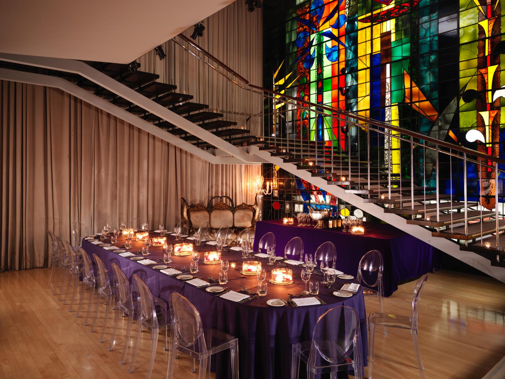 A hotel dining room with a long table, transparent chairs, a lit candelabra, and a stained glass wall at Sanderson London.