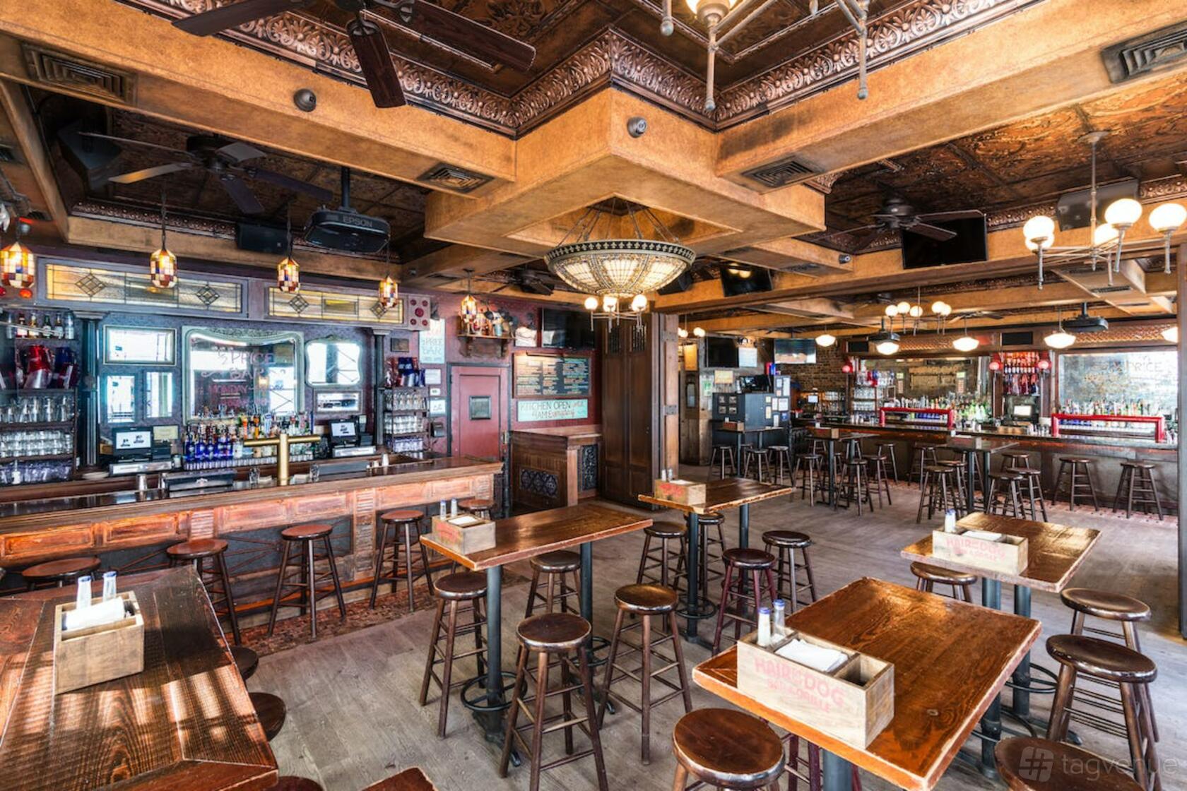 A pub function room with high wooden tables, bar seating, ornate ceiling panels, and pendant lighting at Hair of the Dog.