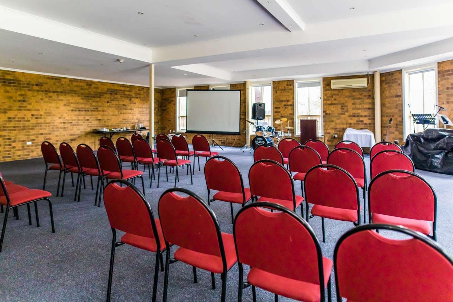 A meeting room with rows of red chairs, brick walls, and a projector screen at Best Western Mill Park Motel.