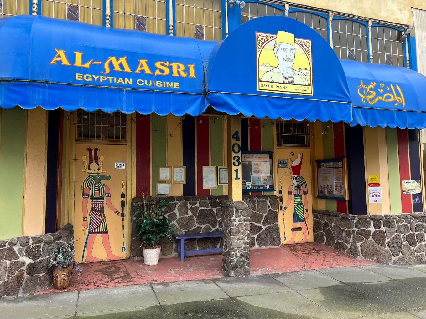 A restaurant exterior with striped walls, Egyptian-themed murals on doors, and a blue awning at Al-Masri Egyptian Restaurant.