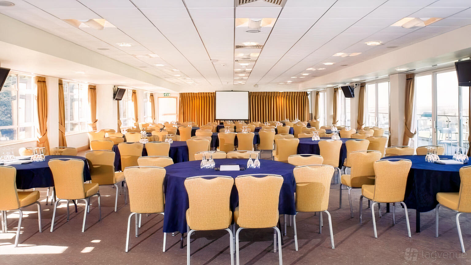 A meeting room with round tables draped in navy linens, tan chairs, and large windows at Sandown Park Racecourse.