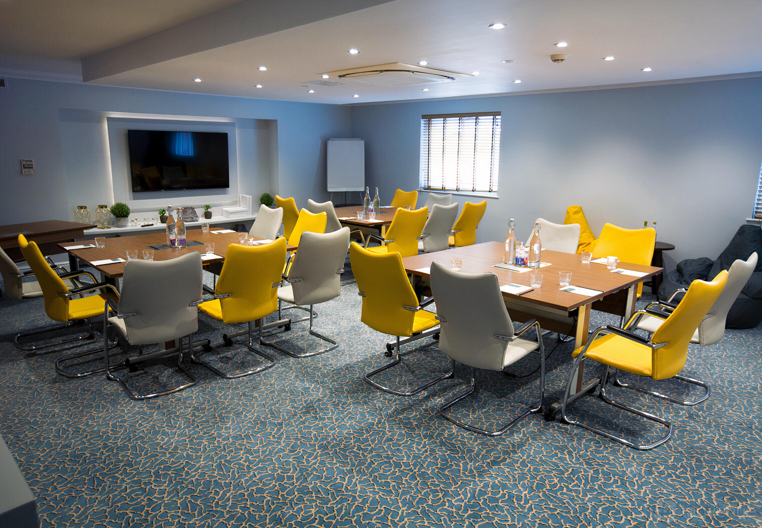 A meeting room with yellow and white chairs around rectangular tables, TV screen, and natural light at Aztec Hotel & Spa.