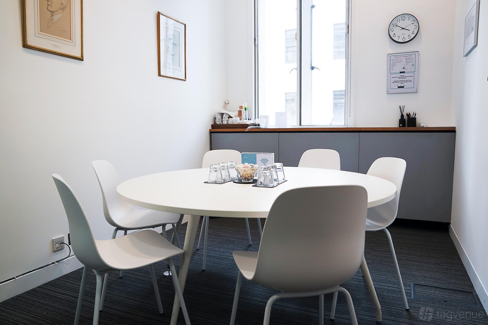 A meeting room with a round table, six white chairs, wall art, and a large window at 1 Wimpole Street.