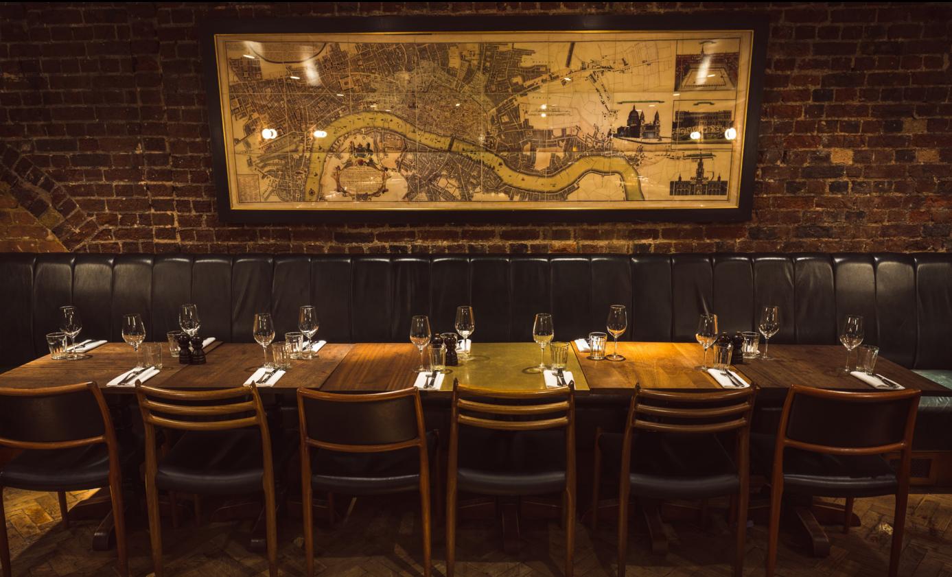 A restaurant with exposed brick walls, banquet seating, and a long table set for group dining at Hawksmoor Spitalfields.