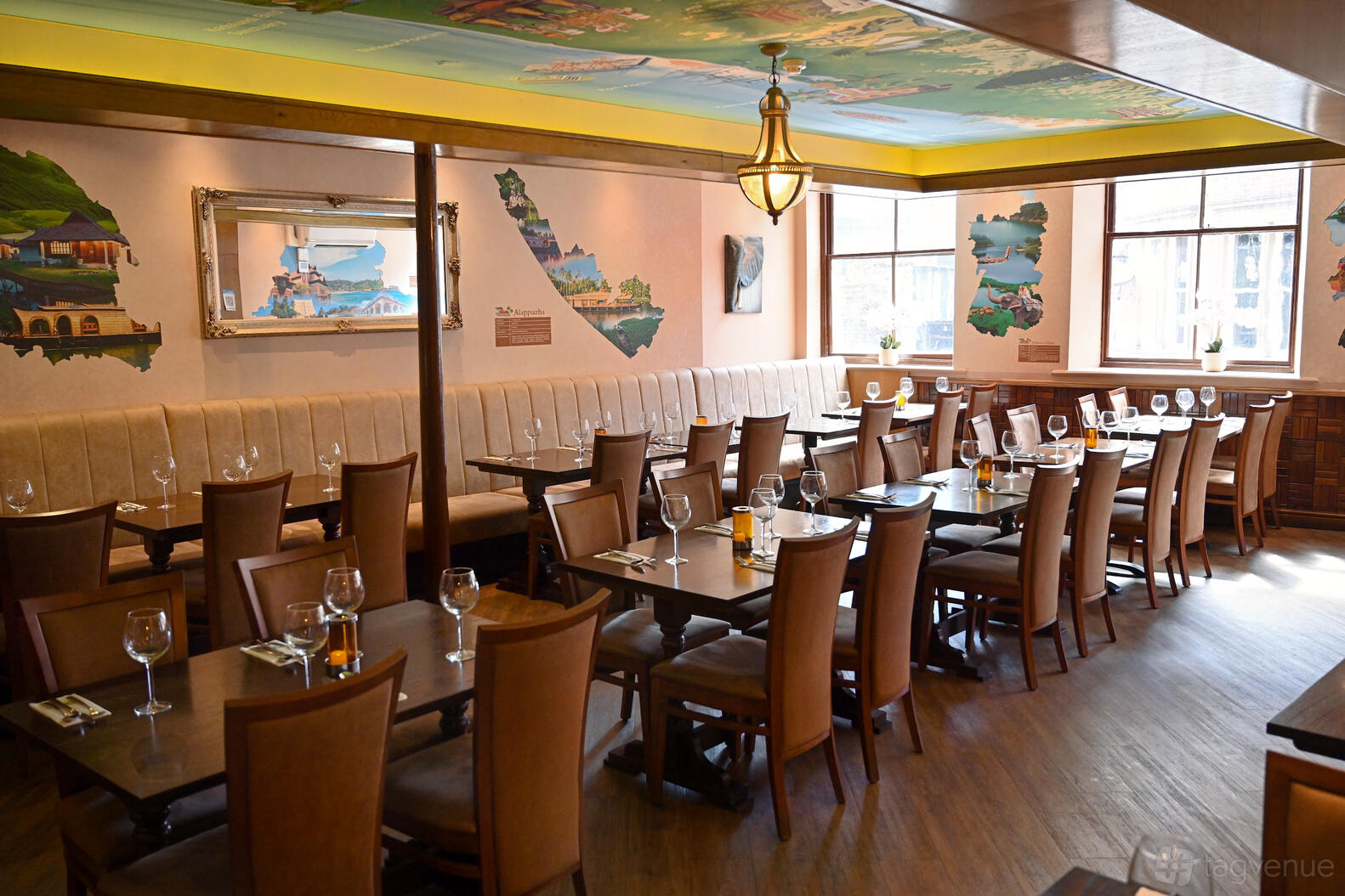An event space in a restaurant with wall art, wooden chairs, and set tables with glassware at Tharavadu.