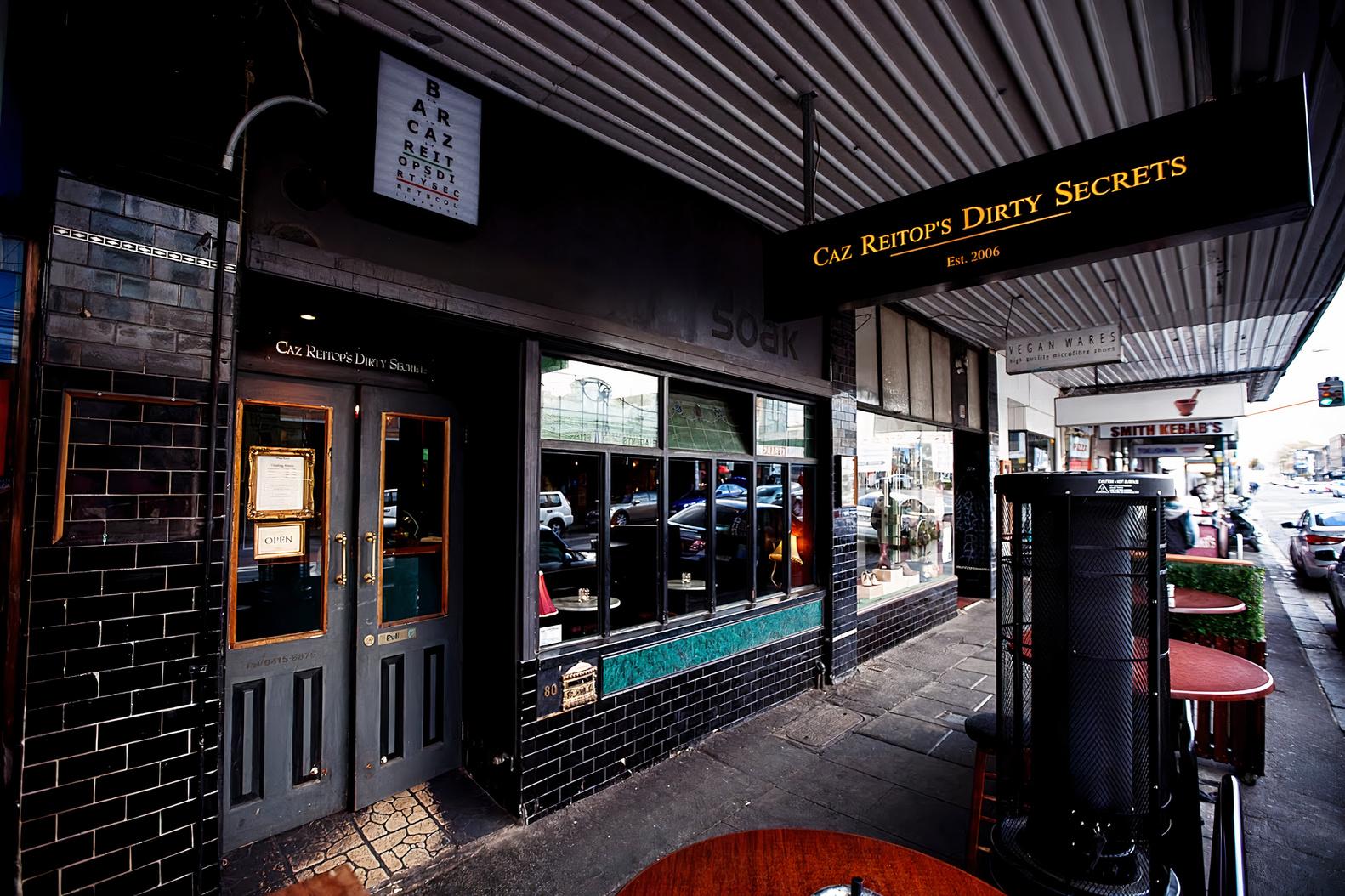 A pub with a black-tiled exterior, large windows, and outdoor tables on the sidewalk at Caz Reitop's Dirty Secrets.