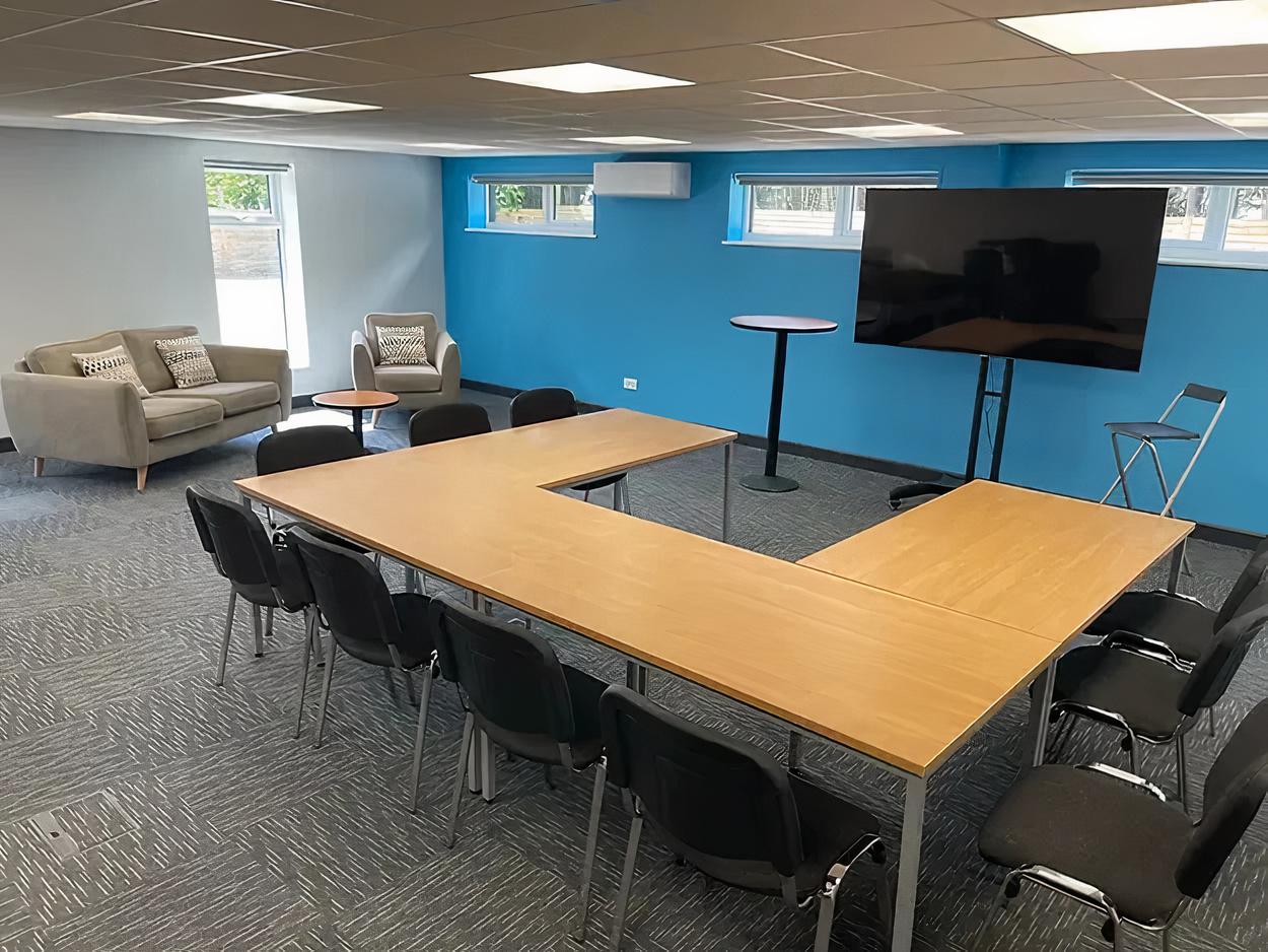 A meeting room with U-shaped tables, black chairs, a wall-mounted screen, and blue accent wall at Crown Hall Farm.
