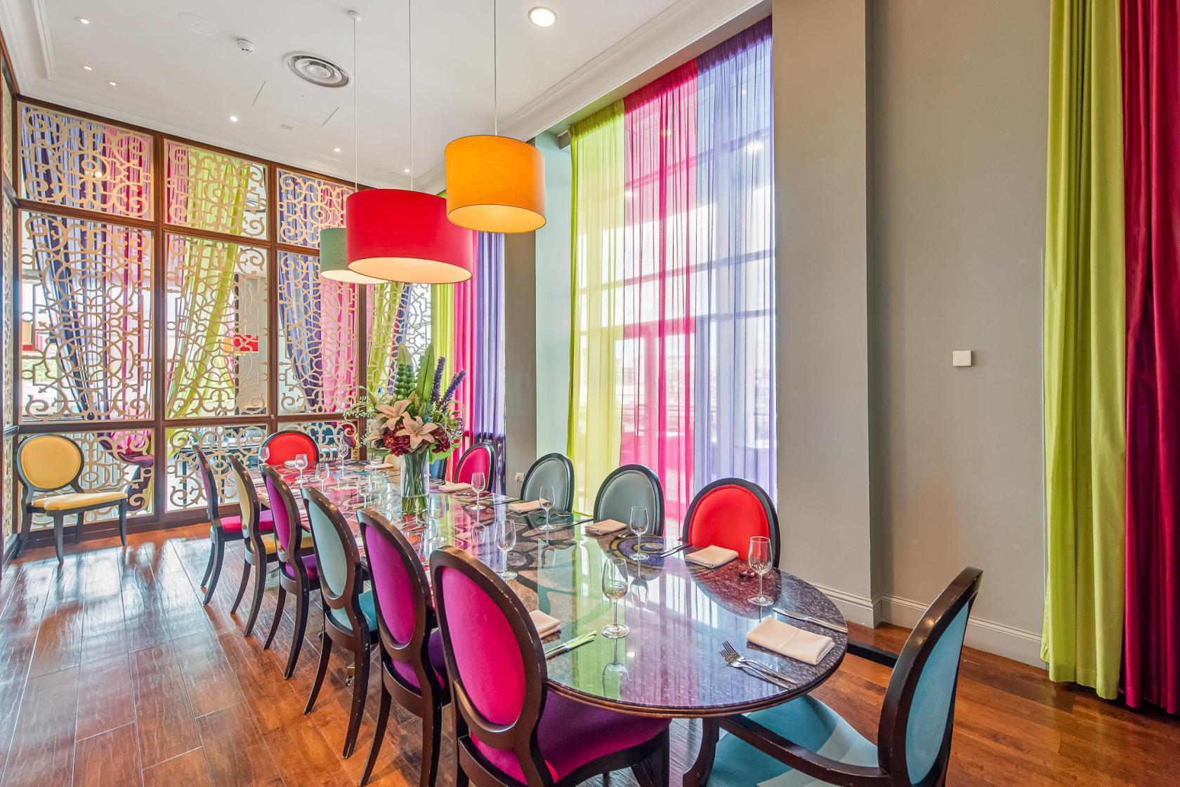 A dining room with a long oval table, colorful chairs, pendant lights, and sheer multicolored curtains at Alea Casino Glasgow.