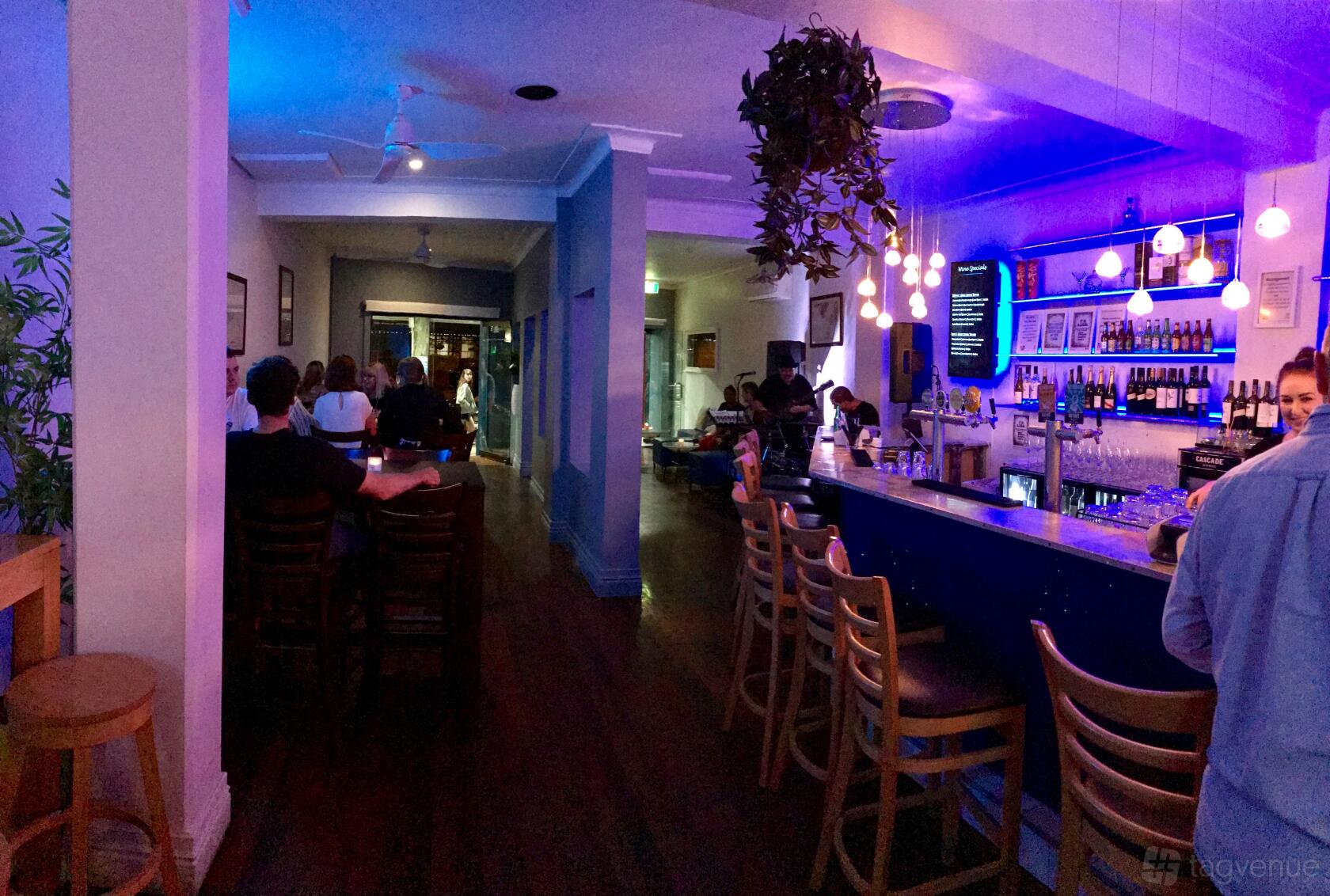 A lounge bar with blue accent lighting, high-top wooden chairs, and a hanging plant at BlueBottle Cocktail & Tapas Bar.