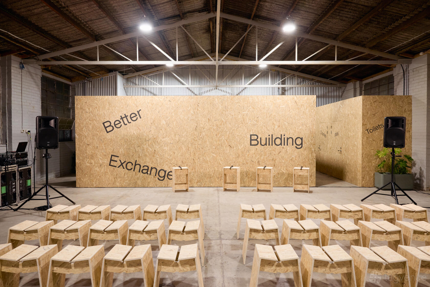 A warehouse event space with plywood walls, exposed beams, wooden stools, and a speaker setup at Better Building Exchange Brunswick.