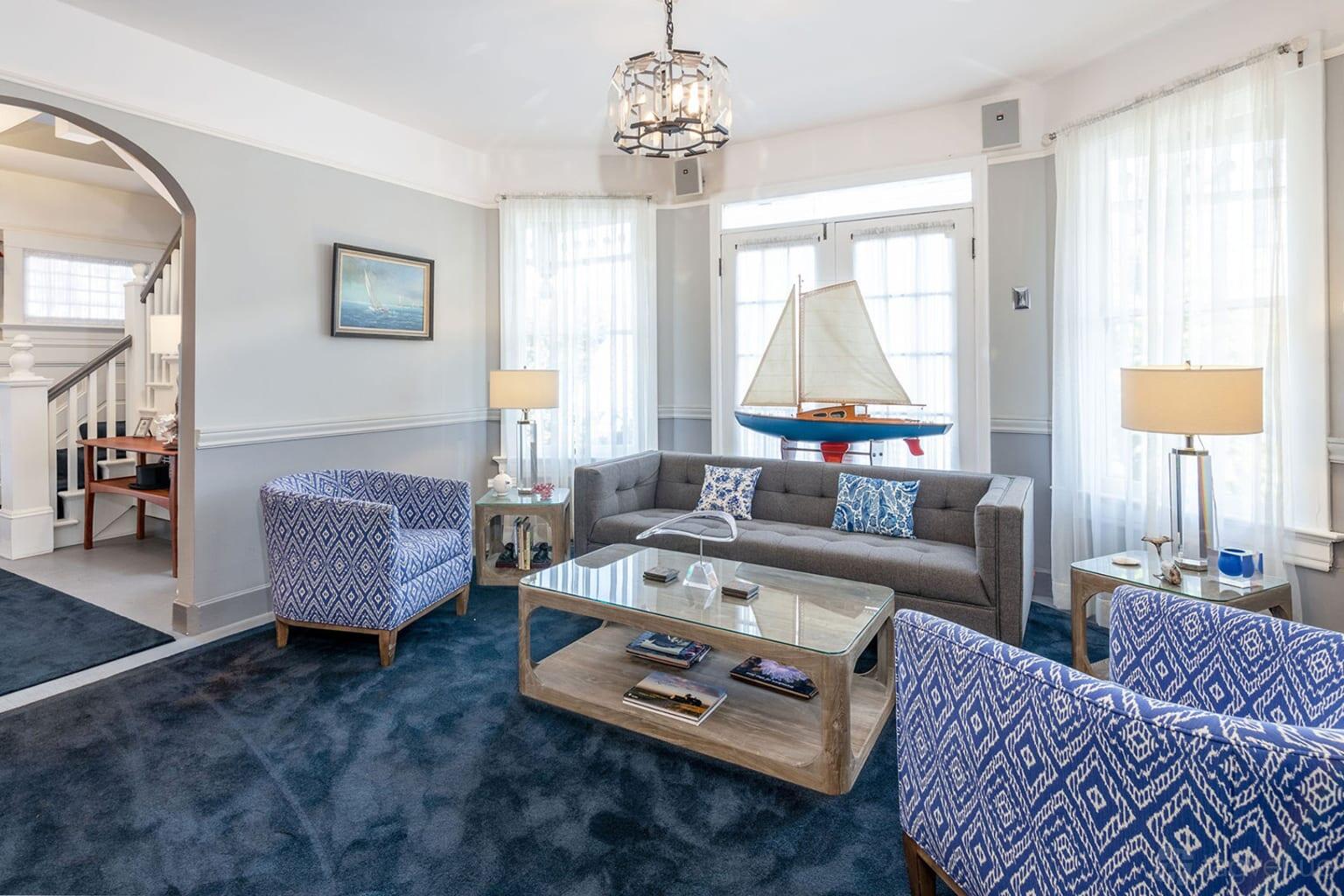 A hotel lounge with patterned blue armchairs, a gray sofa, and a sailboat model by sunlit windows at Nantucket Whale Inn.