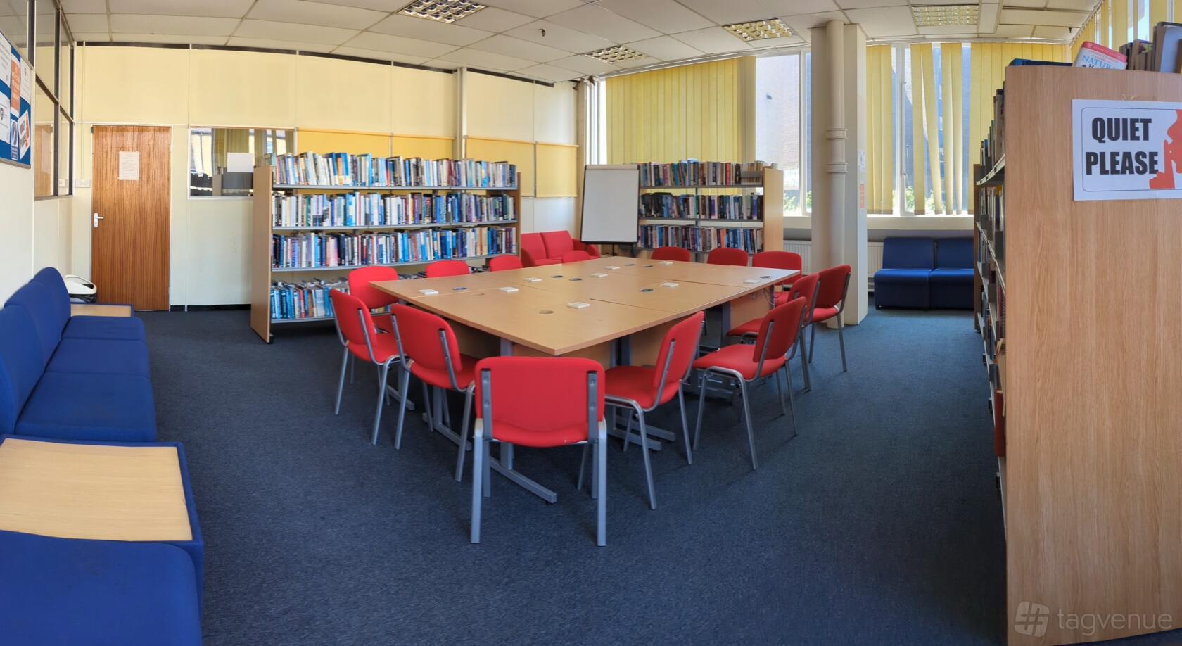 A university library with bookshelves, a large rectangular table surrounded by red chairs, and natural light at The Woolwich College.