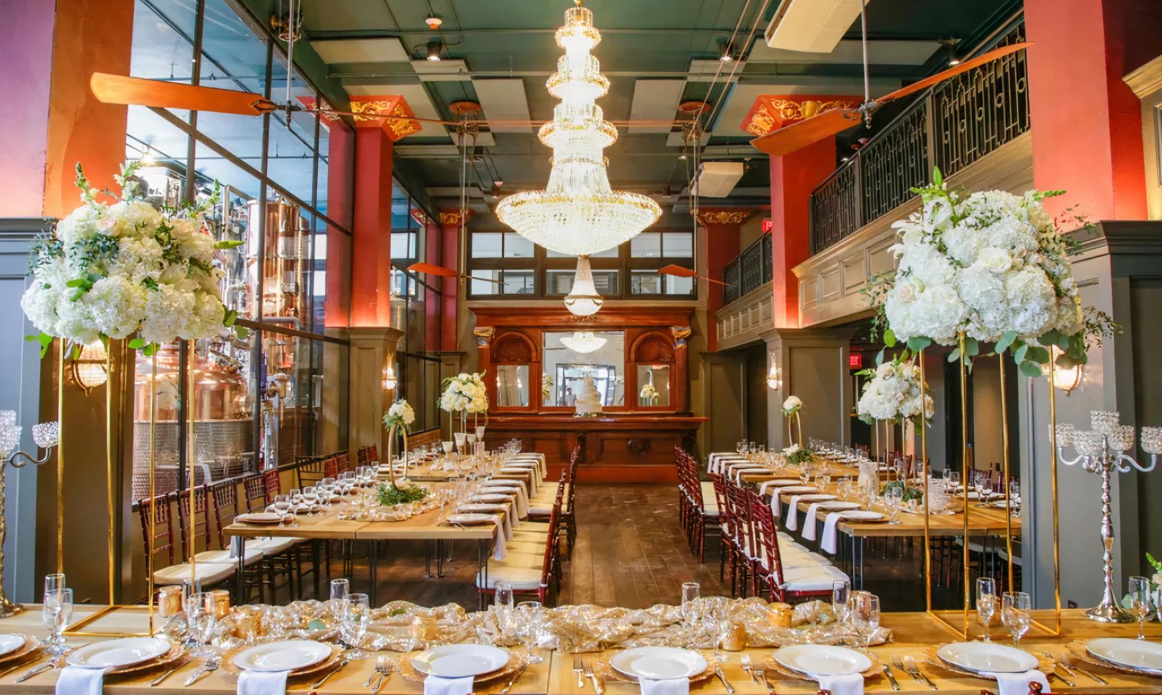 A distillery venue with chandeliers, floor-to-ceiling windows, copper stills, and banquet tables with floral centerpieces at Devils River Distillery.