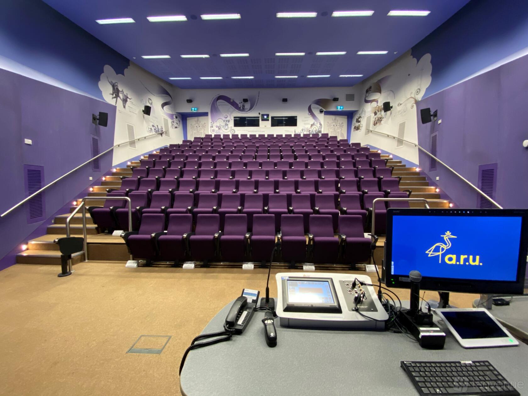 An auditorium with tiered purple seating, wall illustrations, and a presentation control desk at ARU Venue Hire - Chelmsford.