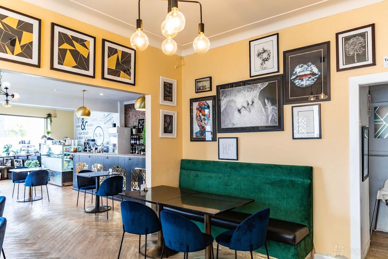 A dining room with velvet green banquette seating, dark tables, and framed artwork on the walls at The Den Cafe and Bar.