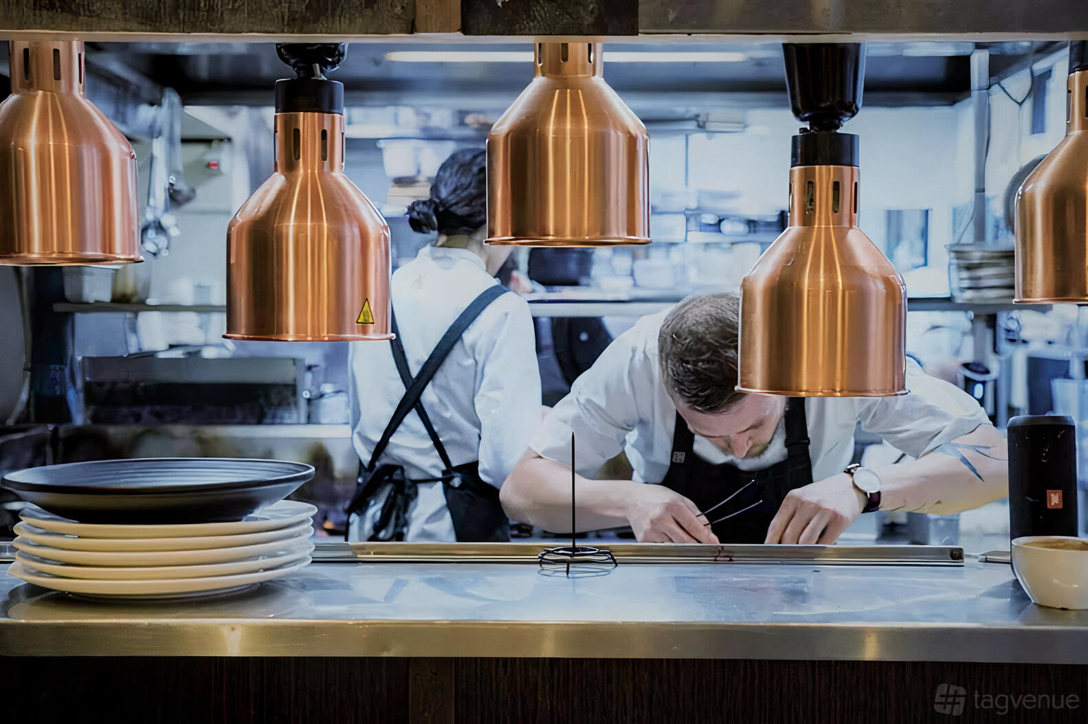 A restaurant kitchen with copper pendant lights, stacks of white plates, and chefs preparing dishes at The French Brasserie.
