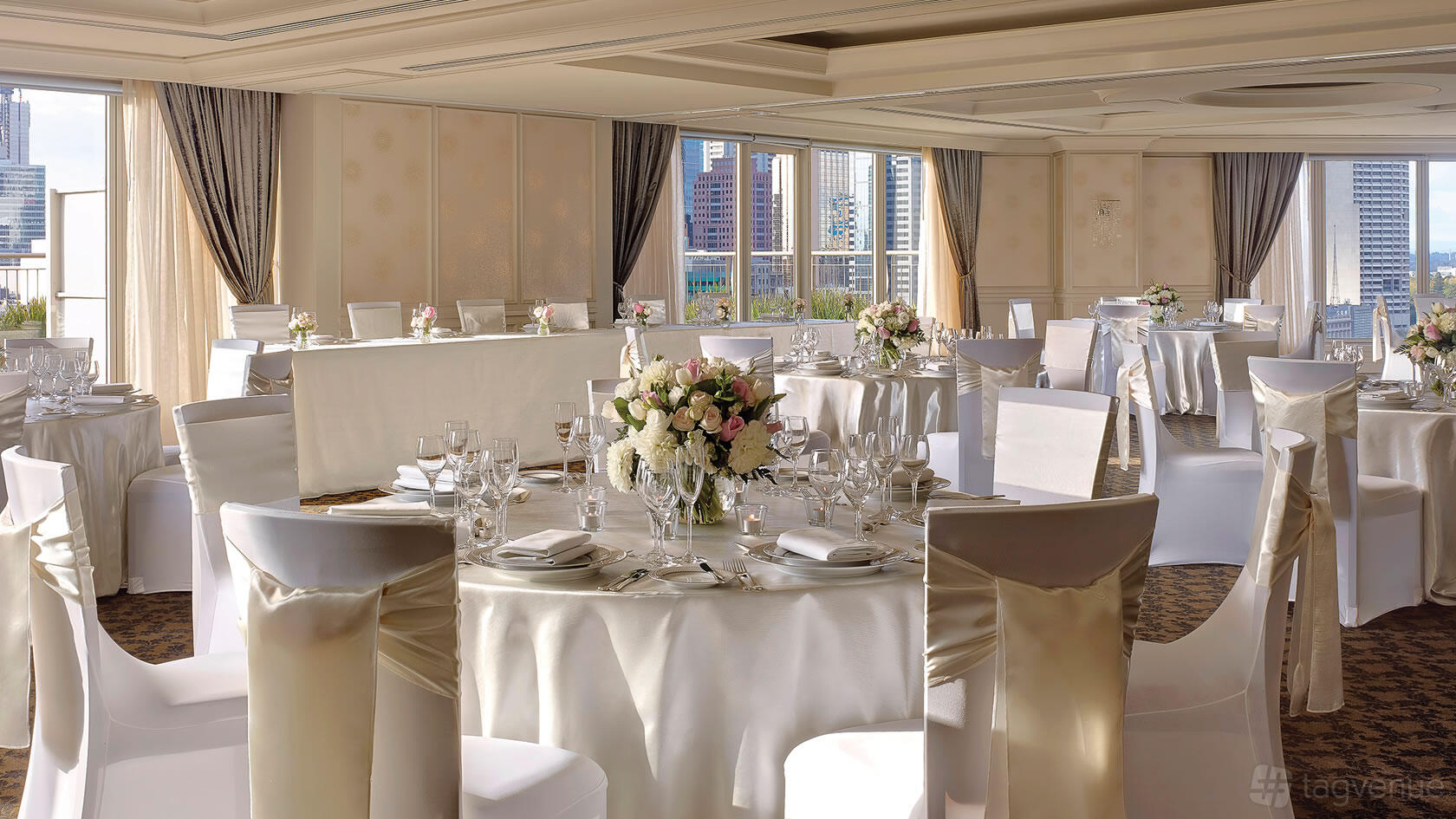 A function room with round tables draped in white linens, floral centerpieces, and city views at The Langham Melbourne.