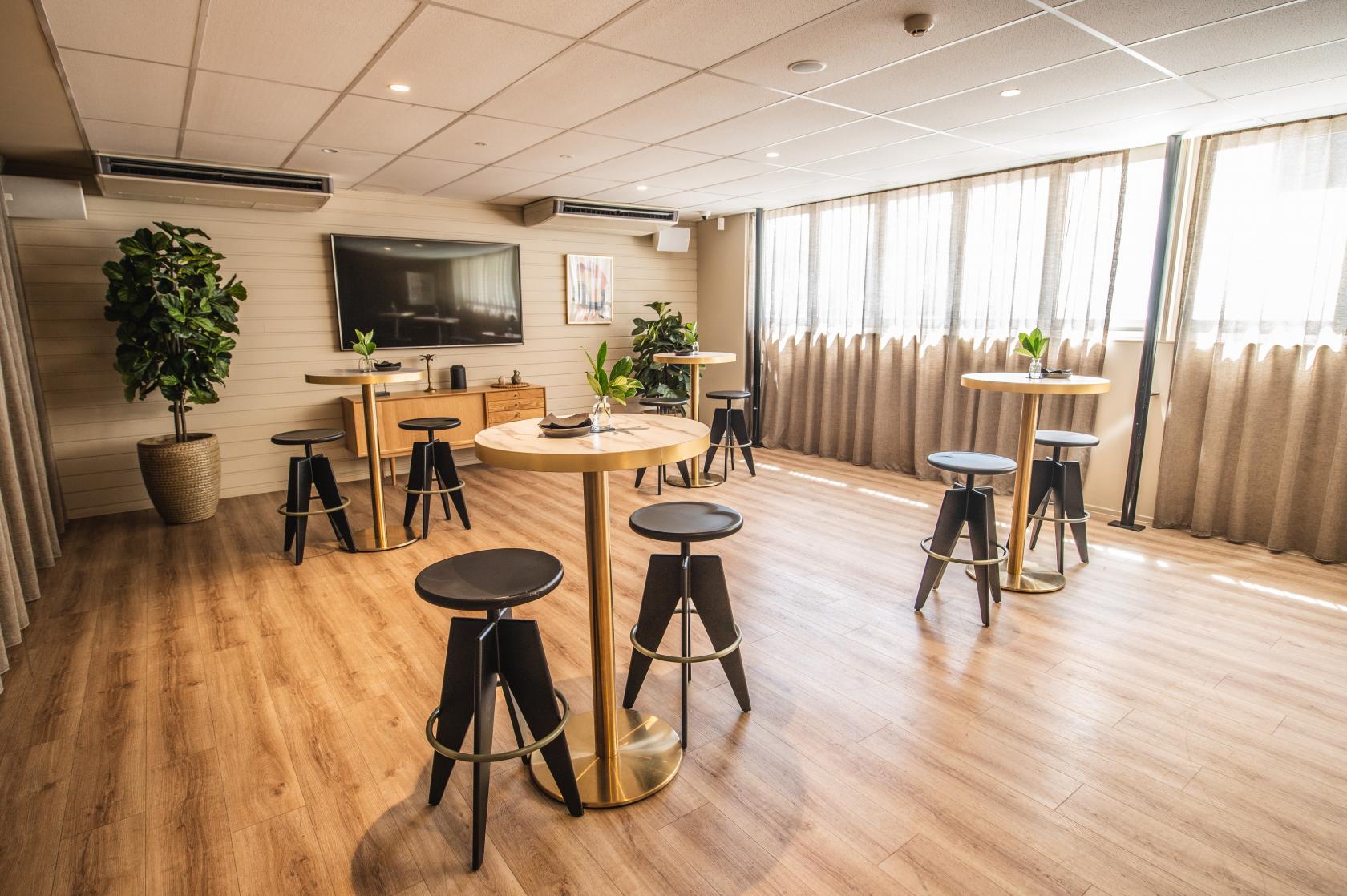 A meeting room with floor-to-ceiling windows, tall round tables, bar stools, and potted plants at The Alcott.