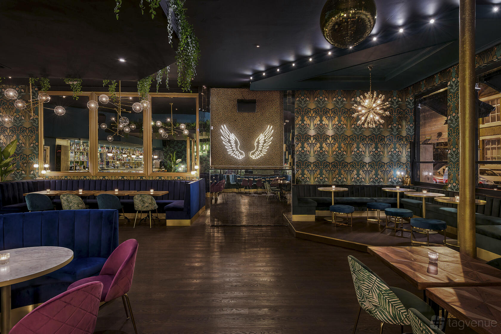 A cocktail bar with velvet seating, patterned wallpaper, globe pendant lights, and a neon wing sign at Lost Society (Battersea SW11).