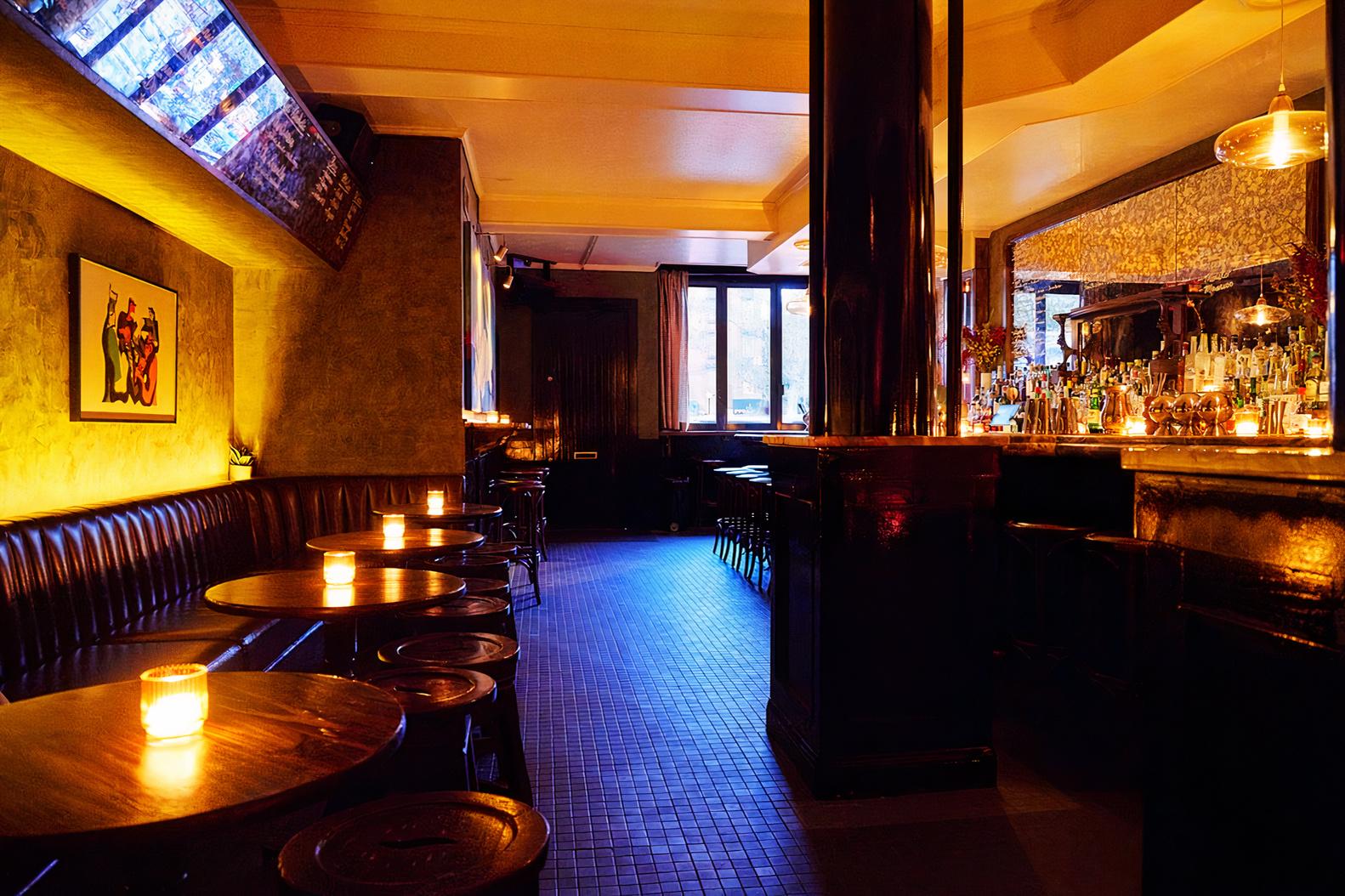 A dimly lit bar with leather banquettes, small round tables, and a long mirrored bar at Two Doors Down.