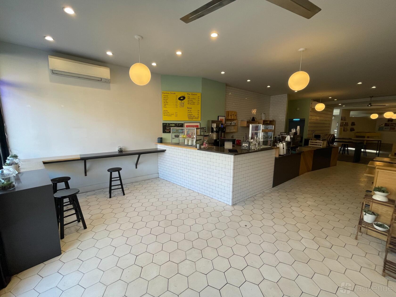 A cafe with a white tiled counter, hexagon tile flooring, globe pendant lights, and window seating at For The Record.
