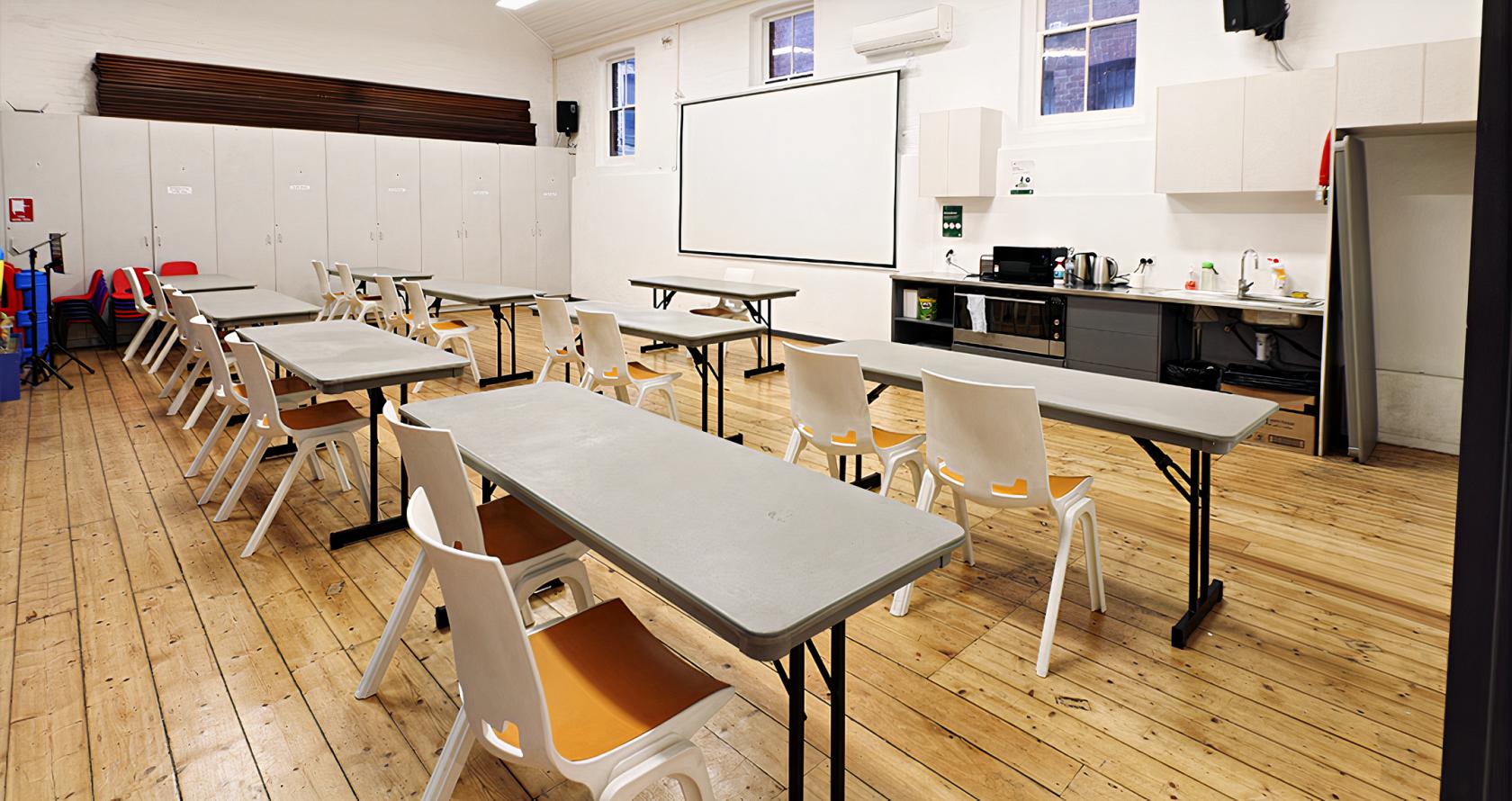 A conference hall with rows of gray tables, white chairs, a large projector screen, and a kitchenette at Melbourne City Conference Centre.