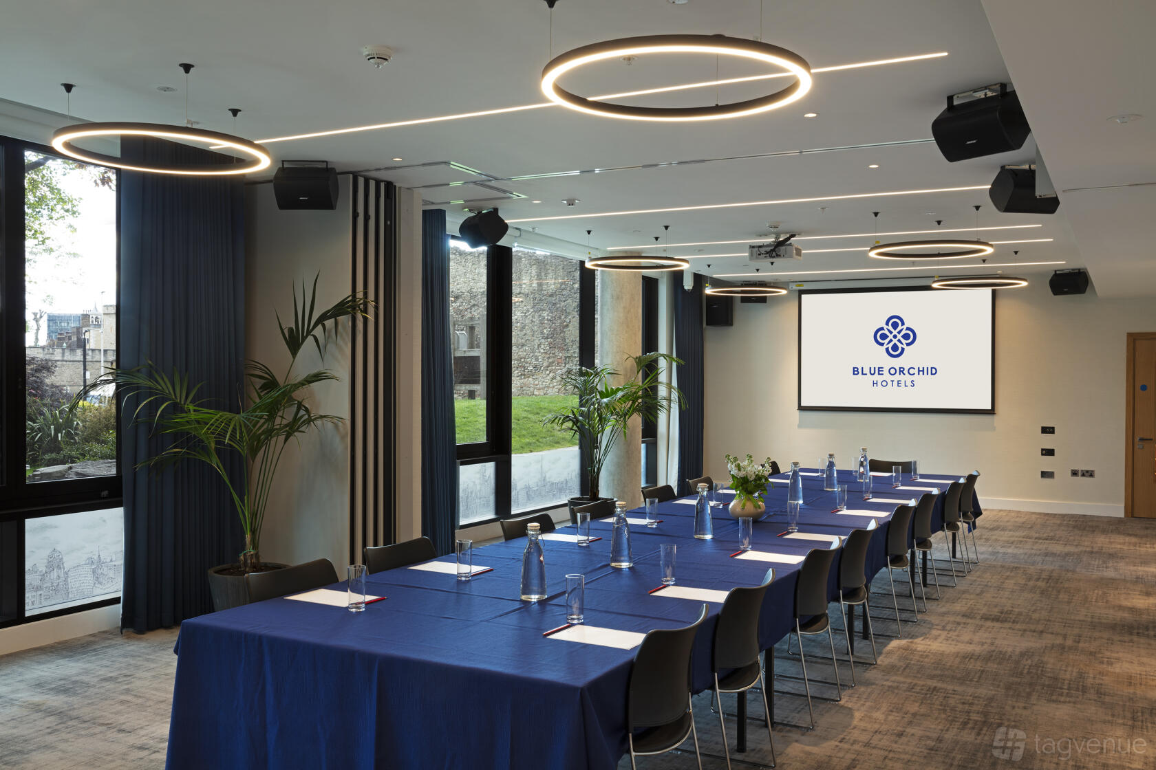 An event space with floor-to-ceiling windows, circular pendant lights, and tables with navy linens at Tower Suites by Blue Orchid.