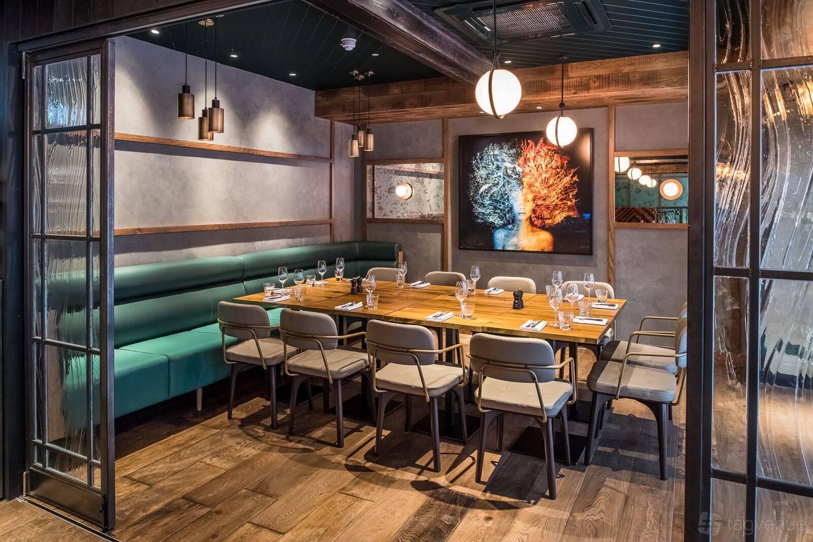 A dining room with a long wooden table, teal banquette seating, pendant lighting, and contemporary art at The Coal Shed- London.