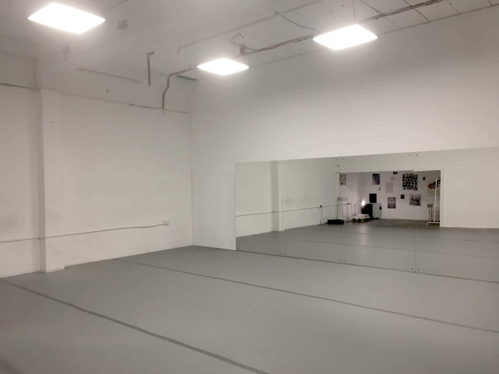 A blank canvas studio with white walls, large wall mirror, and light grey flooring at Ample Space - Hackney Wick at Wallis Road.