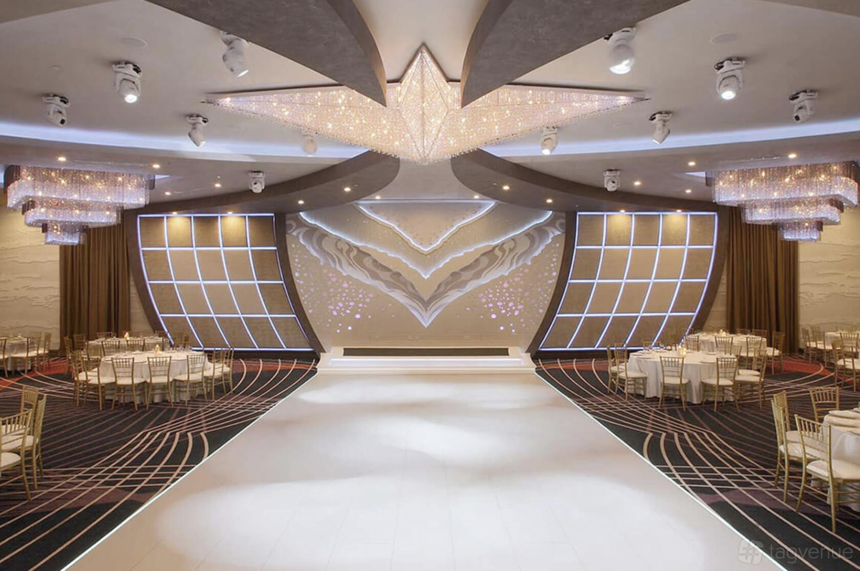 A ballroom with a large illuminated stage, star-shaped crystal chandelier, round tables, and gold chairs at Allure Banquet & Catering.