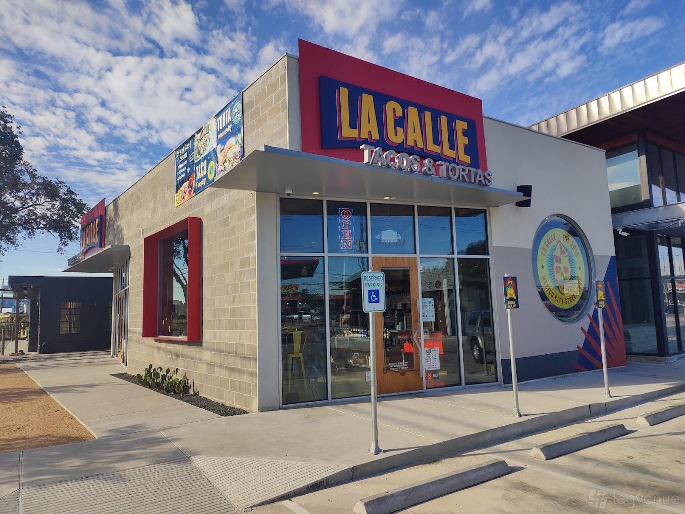 A restaurant with a glass entrance, large signage, and shaded sidewalk seating at La Calle Ella.
