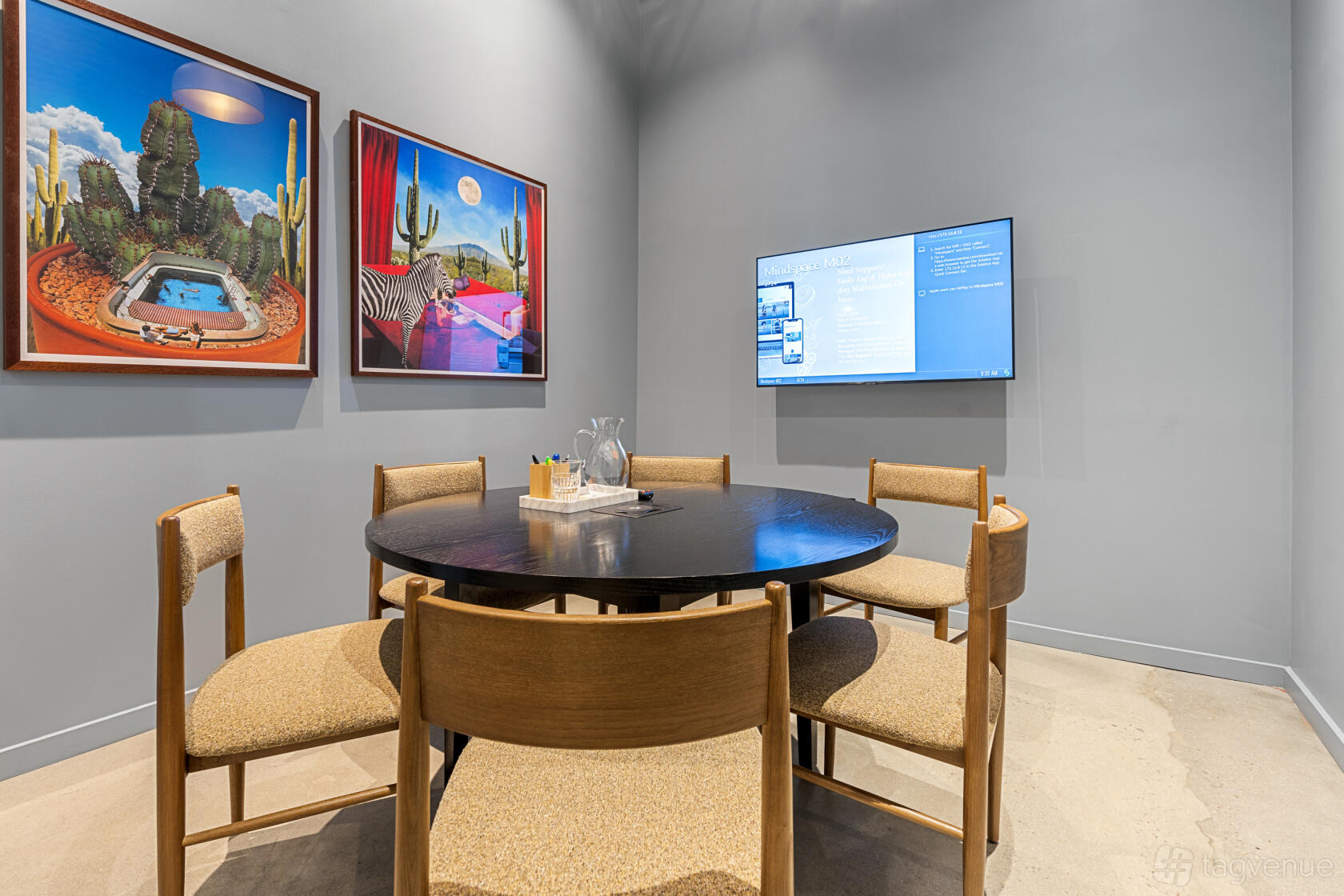 A coworking meeting room with a round table, five chairs, wall art, and a mounted screen at Williamsburg.