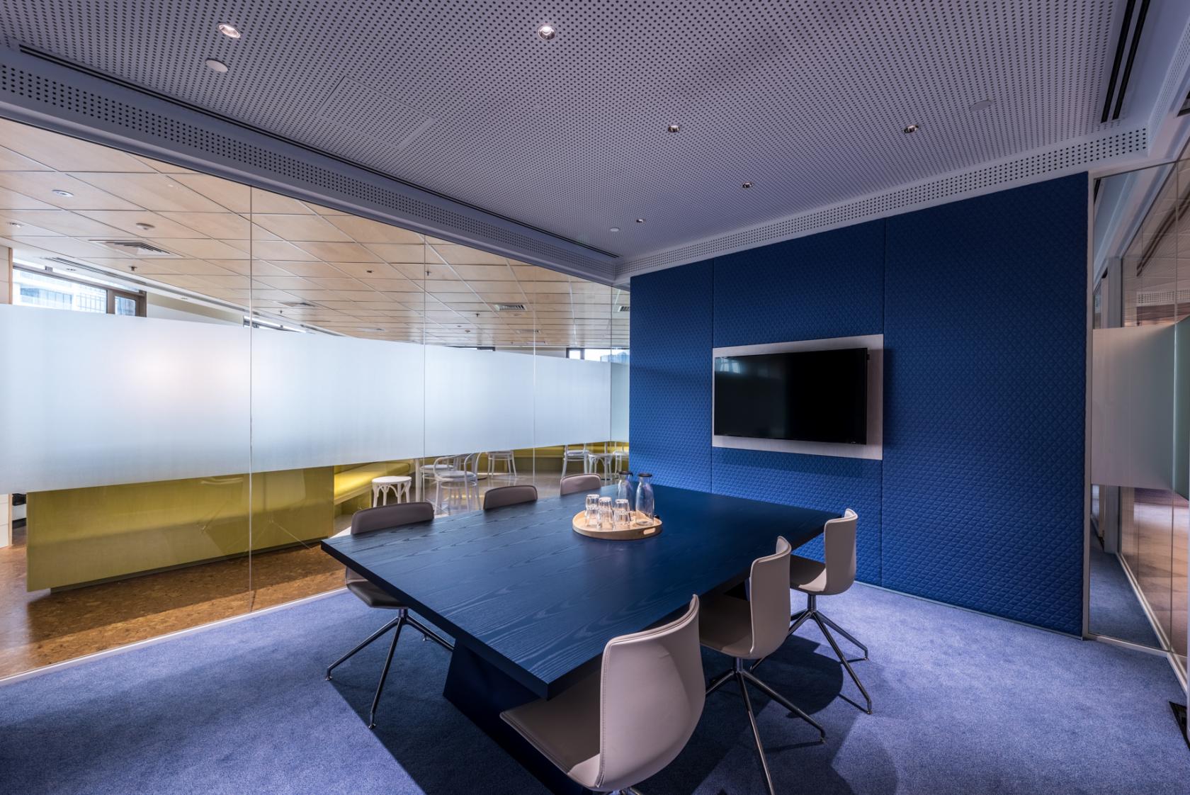 A meeting room with a navy conference table, six gray chairs, frosted glass walls, and a wall-mounted screen at The Work Project Great World City.