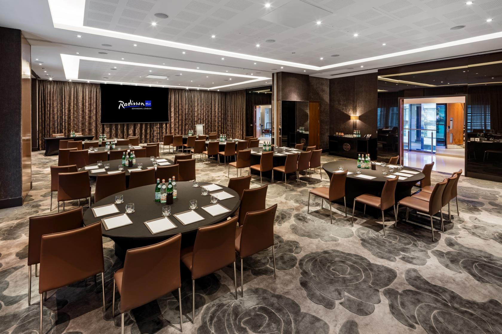 An event space with round and rectangular tables, leather chairs, and a large projection screen at Radisson Blu Hotel London Canary Wharf East.